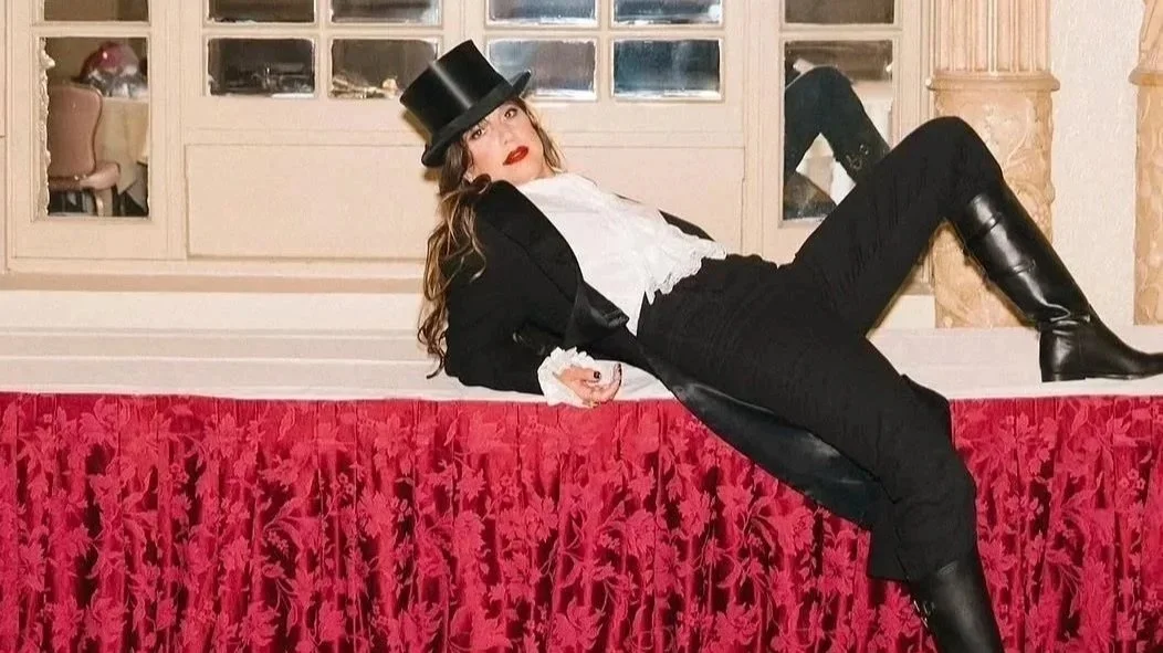 Woman in theatrical costume lying on a red velvet stage, wearing a top hat, black jacket, white shirt, black pants, and tall black boots, with a fancy interior background.