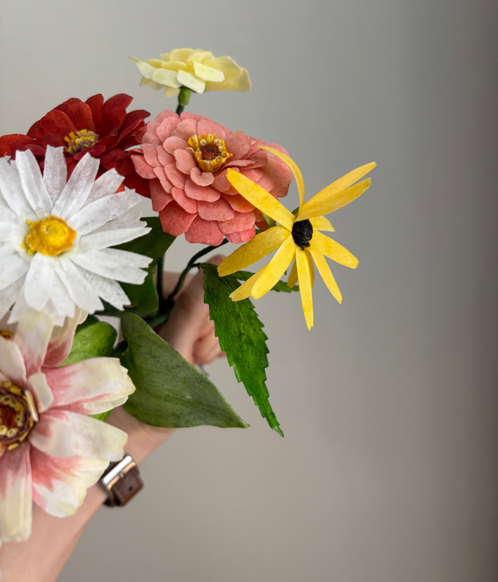 Closeup+of+a+hand+holding+a+bouquet+of+wafer+paper+summer+flowers+including+zinnia%2C+daisy%2C+and+black-eyed+susan.png