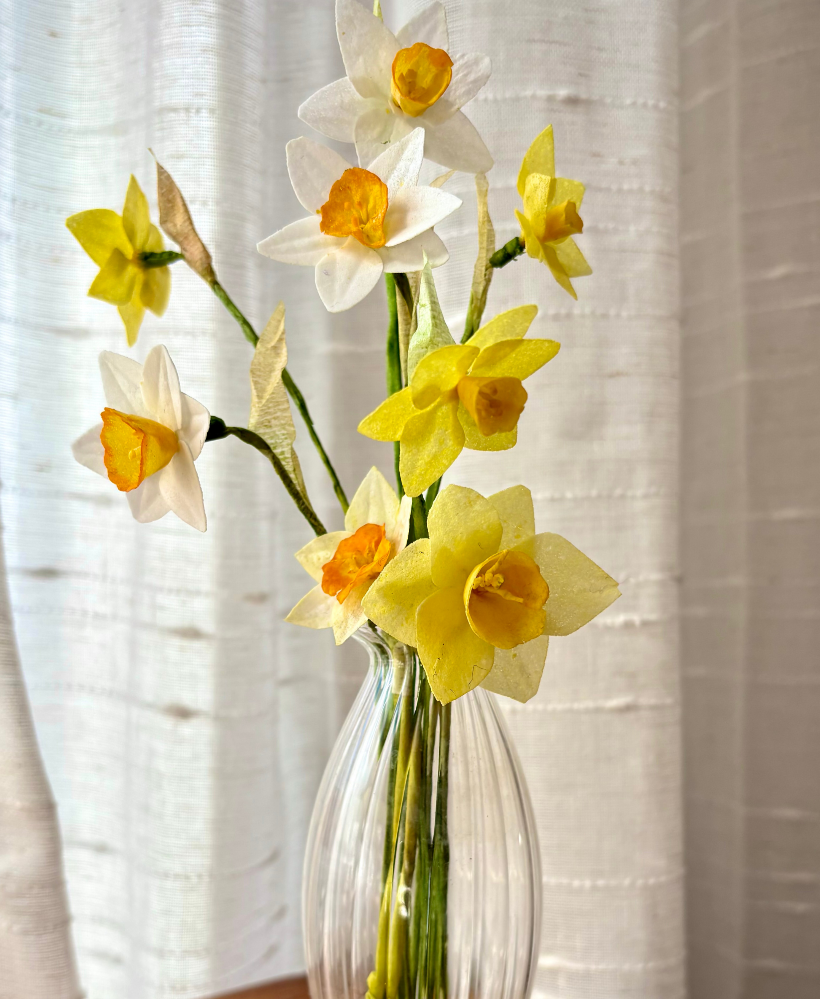 Closeup+of+a+small+fluted+glass+bud+vase+filled+with+wafer+paper+white+and+yellow+and+orange+narcissus+with+a+white+curain+in+the+background.png