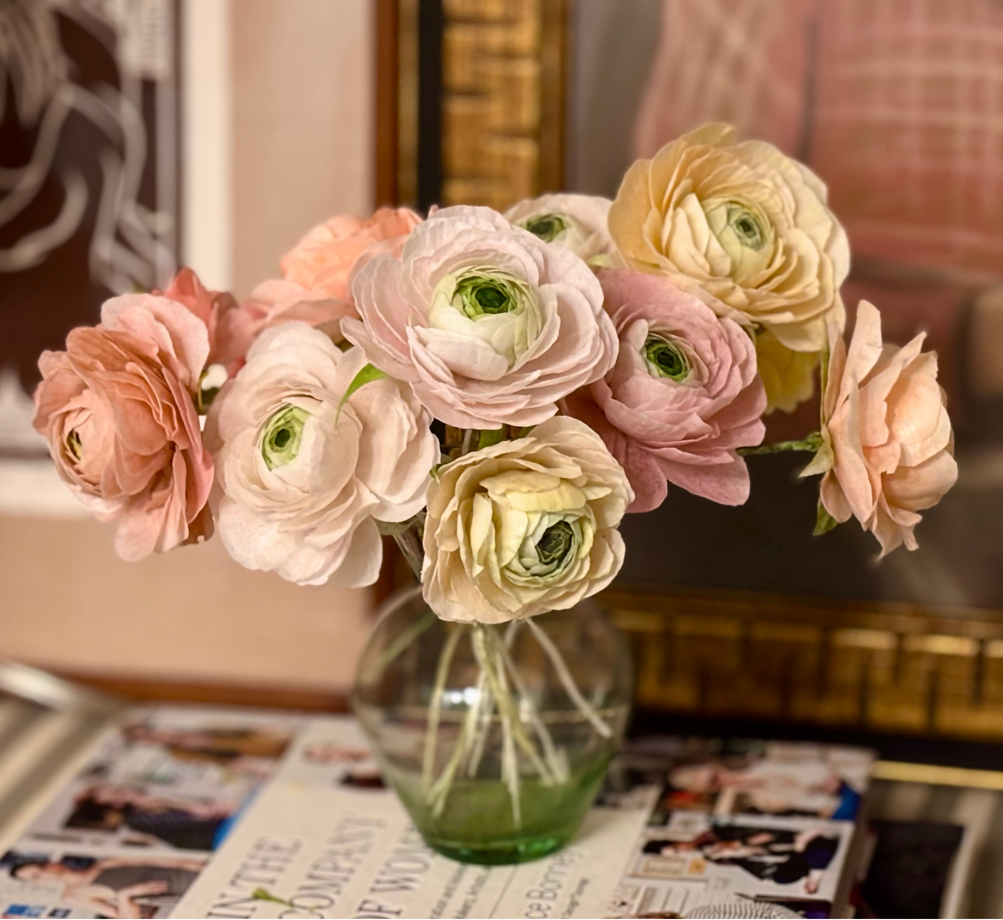 Closeup+of+small+green+glass+budvase+filled+with+an+array+of+muted+peach%2C+yellow%2C+and+pink+wafer+paper+ranunculus%2C+sititng+on+a+coffee+table+with+framed+paintings+in+the+background.png