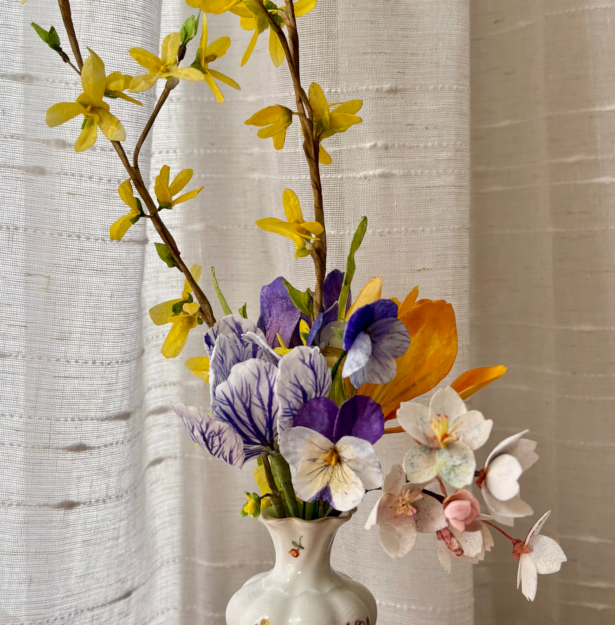 Closeup+of+a+small+ceramic+vase+filled+with+wafer+paper+crocuses%2C+chery+blossoms%2C+violas%2C+and+forsythia%2C+with+a+white+curtain+in+the+background.png