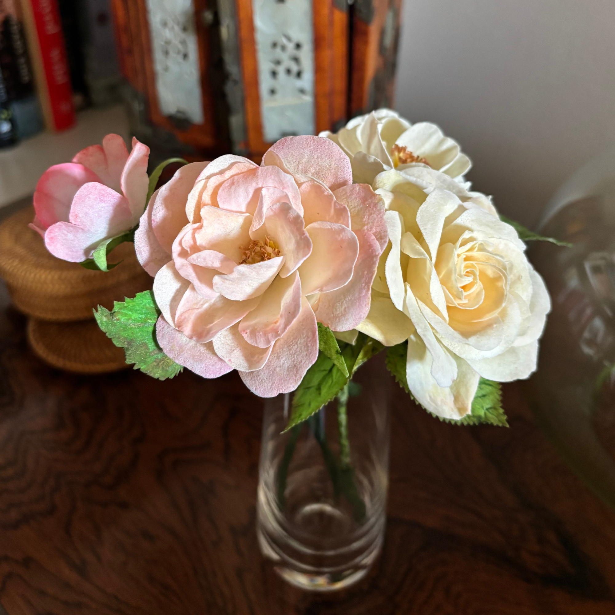 Closeup of small glass vase with a few wafer paper tea roses in blush, pink, cream, and yellow, sitting on a wooden side table with books and knicknacks in the background.png
