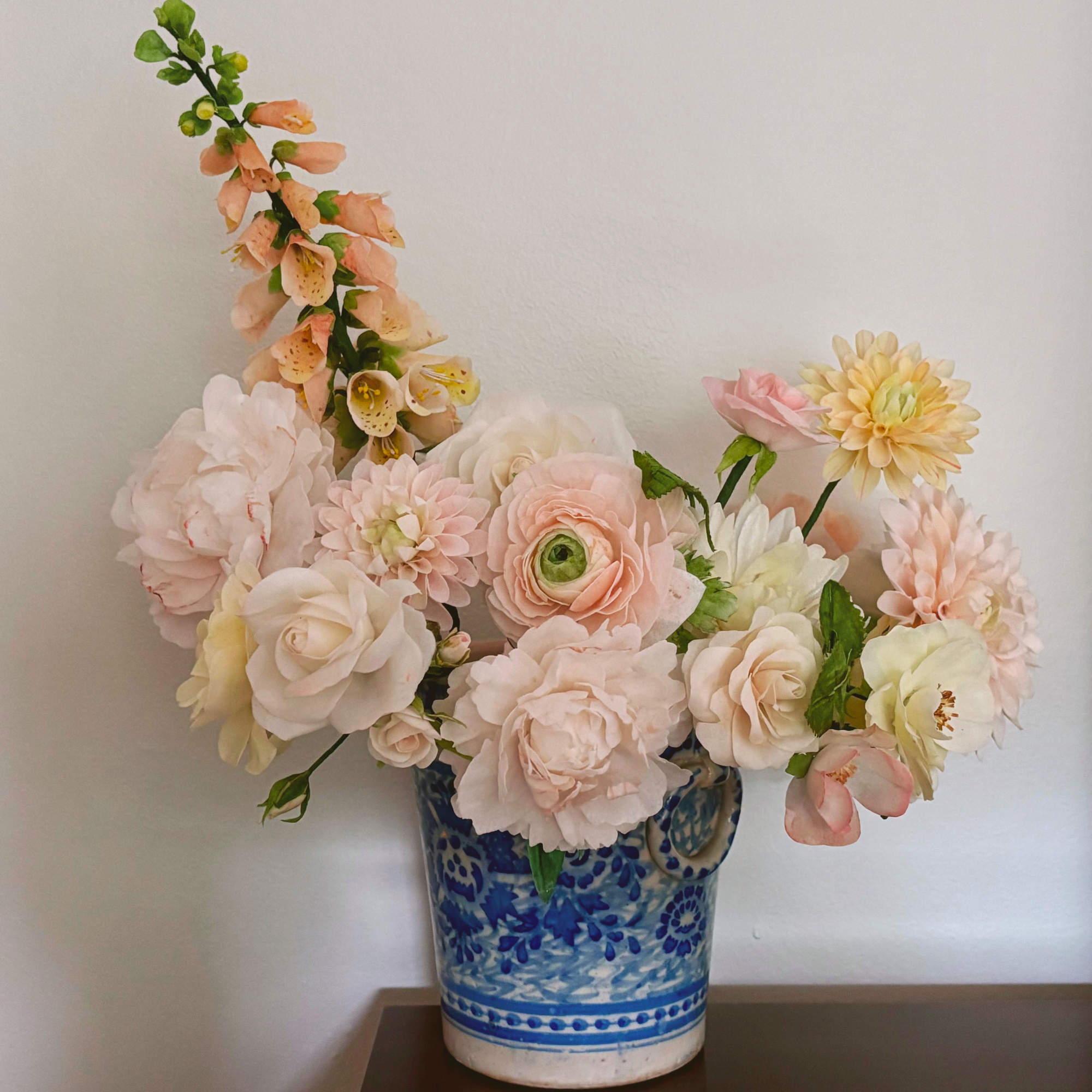 Closeup of a large arrangement of wafer paper flowers, including a peach foxglove, pink roses, white cosmos, peach ranunculus, white peonies, and pink dahlia in blue and white ceramic flower pot.png
