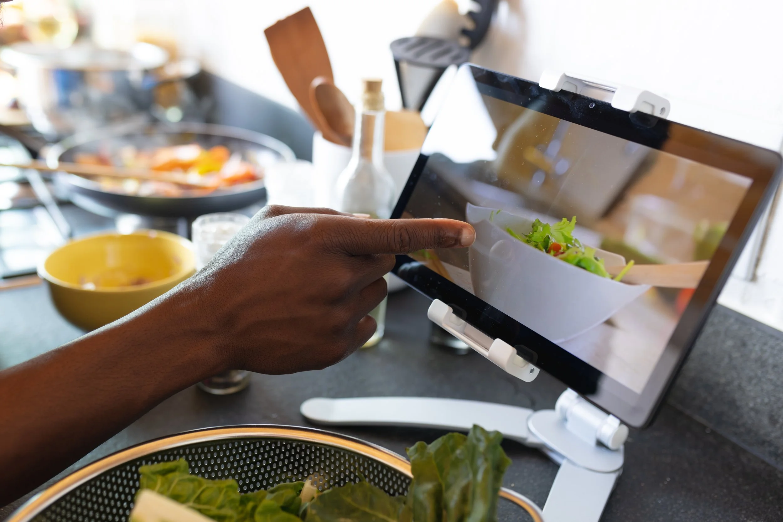 Persona tomando una foto de una ensalada en una tablet en una cocina con varios ingredientes y utensilios en la mesa.
