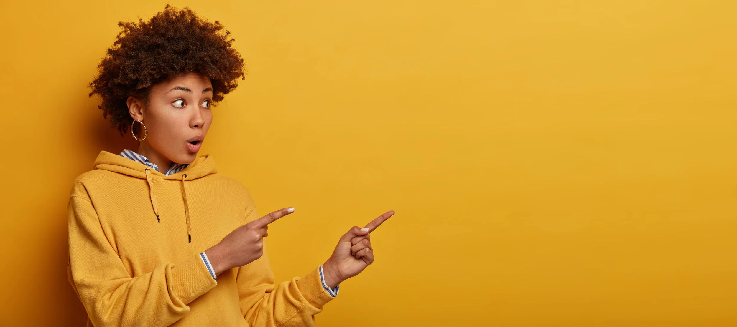 A young woman with curly hair and hoop earrings wearing a yellow hoodie, pointing at the right side with an surprised expression in front of a yellow background.