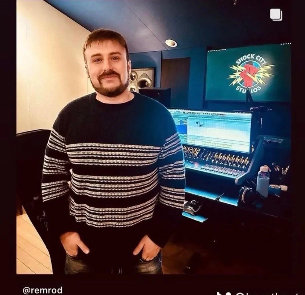A man stands in a recording studio in front of a mixing console and a computer monitor, with a logo for Shock City Studios on a screen behind him.