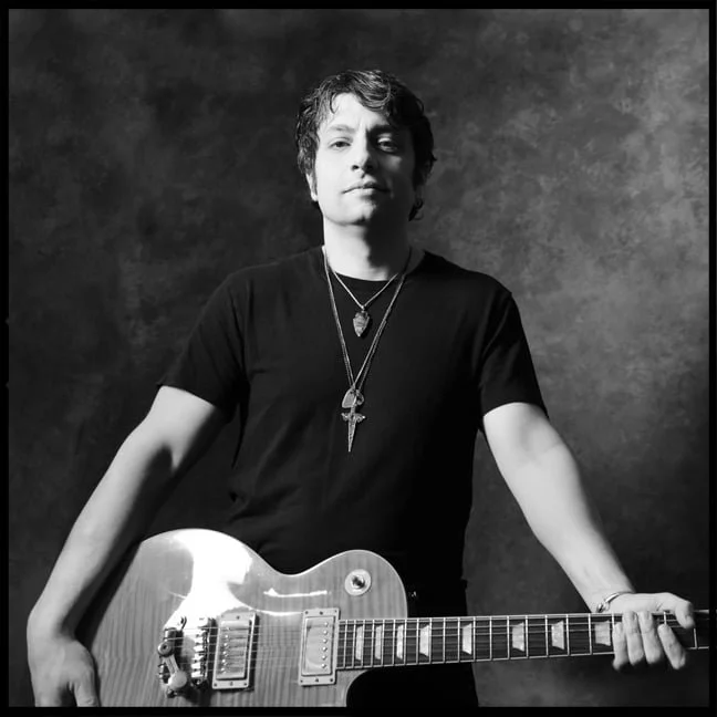 Black and white photo of PETE MURANO with dark wavy hair, wearing a black T-shirt. He has necklaces, including a cross, and is holding an electric guitar, standing against a dark textured background.