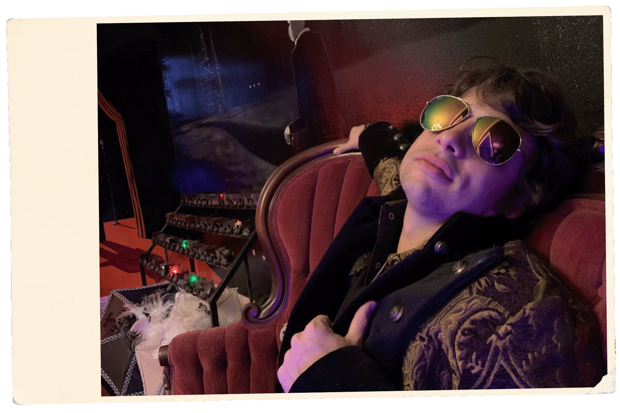 PETE MURANO with sunglasses resting on red velvet chair, wearing a black and gold patterned jacket, in a dimly lit room with red and blue lighting, beside a table with decorative items.