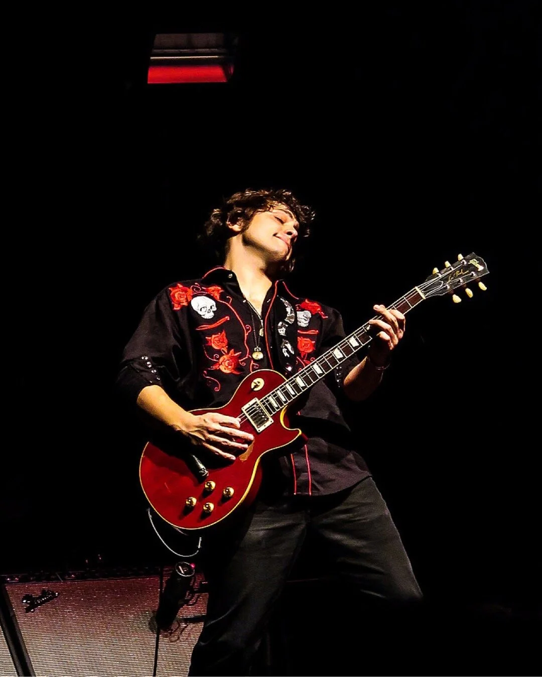 PETE MURANO with dark curly hair playing a red electric guitar on stage, wearing a black embroidered shirt with red accents, against a dark background.