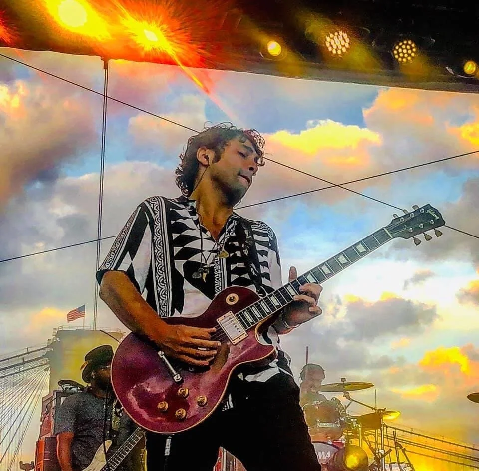 PETE MURANO playing an electric guitar on stage during sunset with colorful clouds in the sky and stage lighting overhead.