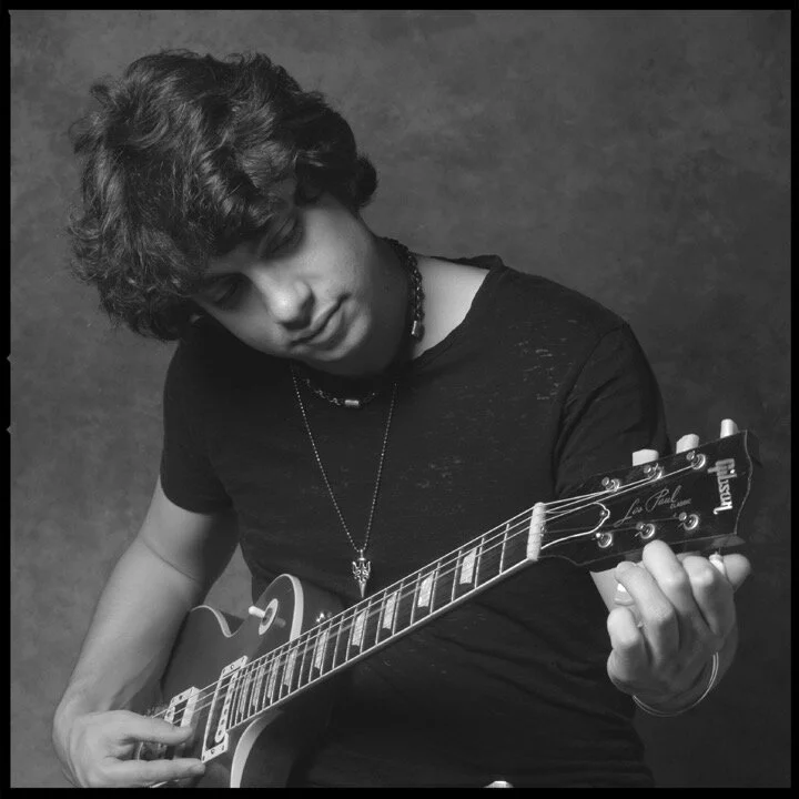 PETE MURANO with curly hair playing an electric guitar, wearing a black shirt and a necklace, against a plain background.