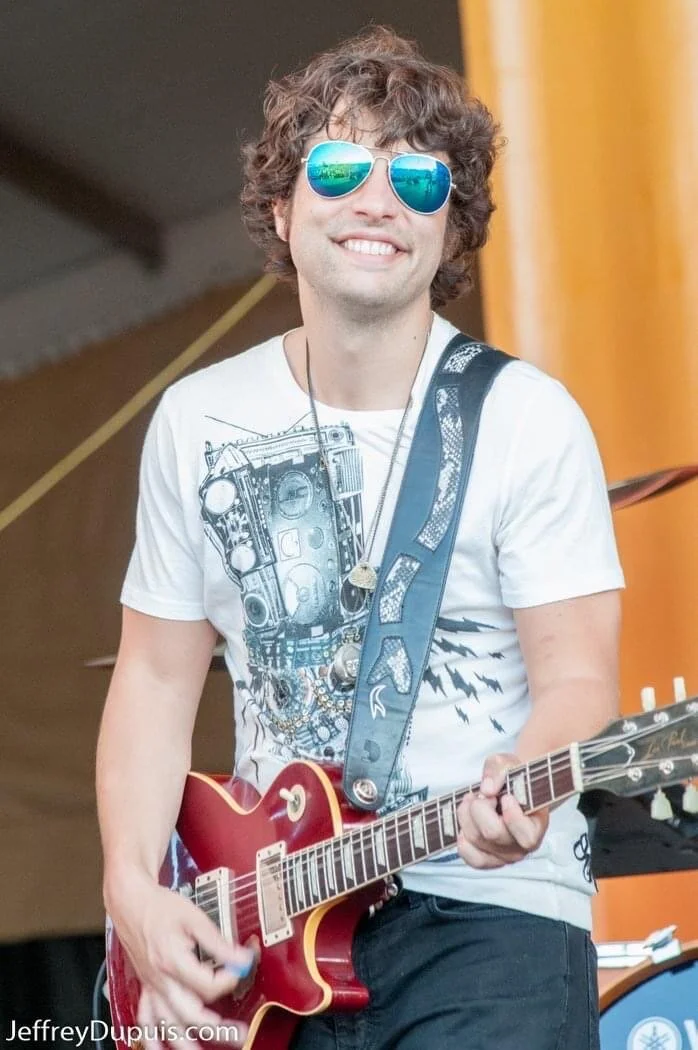 PETE MURANO with curly hair wearing reflective sunglasses, a white graphic t-shirt, and a necklace, smiling while playing a red electric guitar on stage.