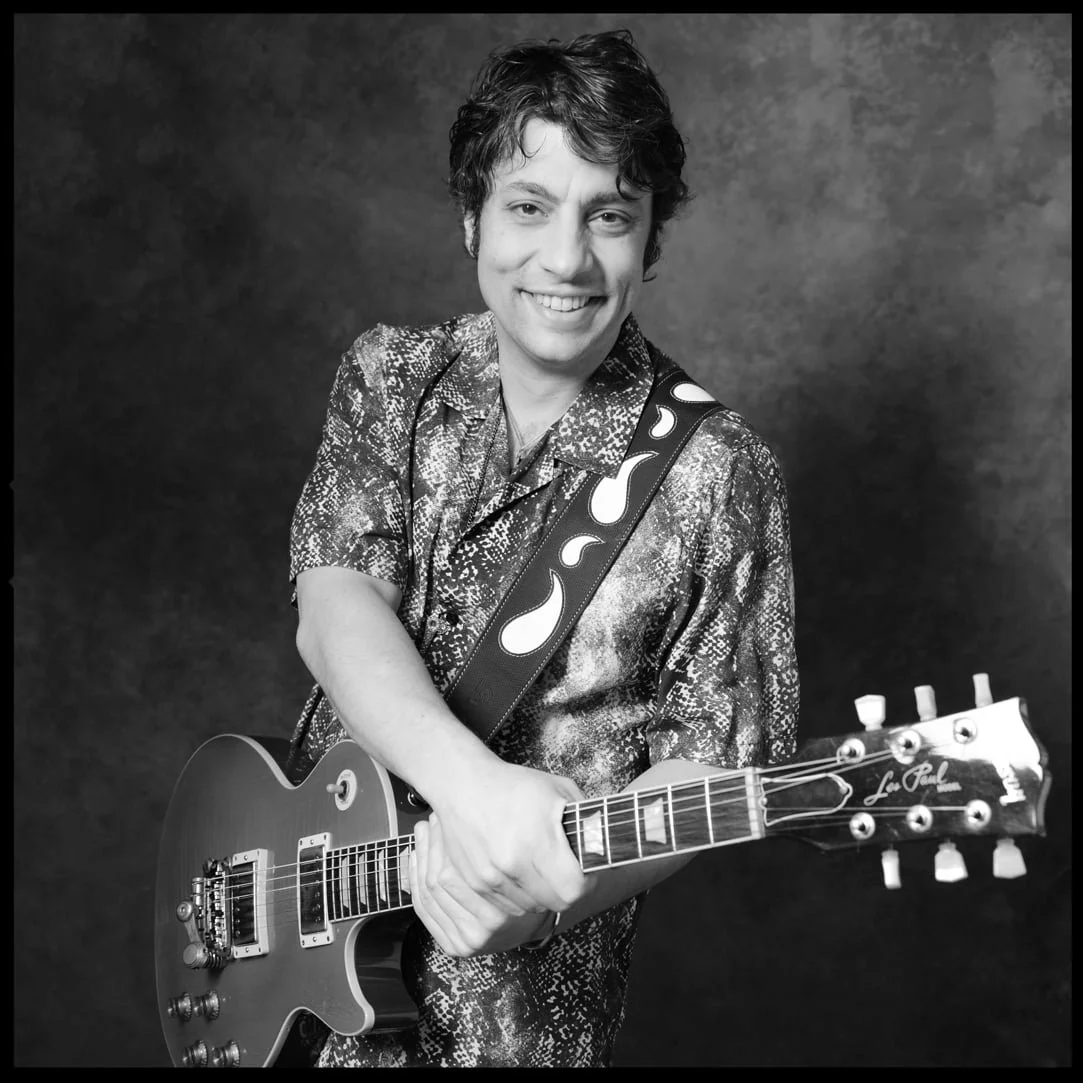 Black and white photo of a young man with curly hair smiling and holding an electric guitar. He is wearing a shiny, patterned shirt and standing against a dark background.
