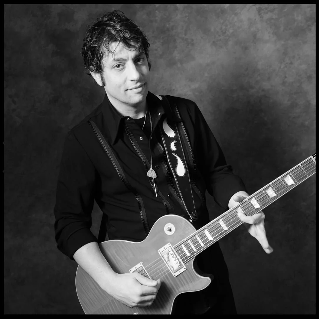 Black and white portrait of PETE MURANO with wavy hair playing an electric guitar, wearing a dark shirt and a guitar strap with a flame pattern, standing against a textured background.