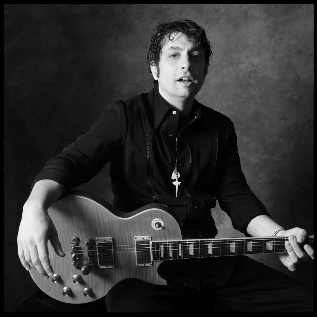 Black and white photo of PETE MURANO sitting and playing an electric guitar, wearing a dark shirt and a necklace with a cross pendant.