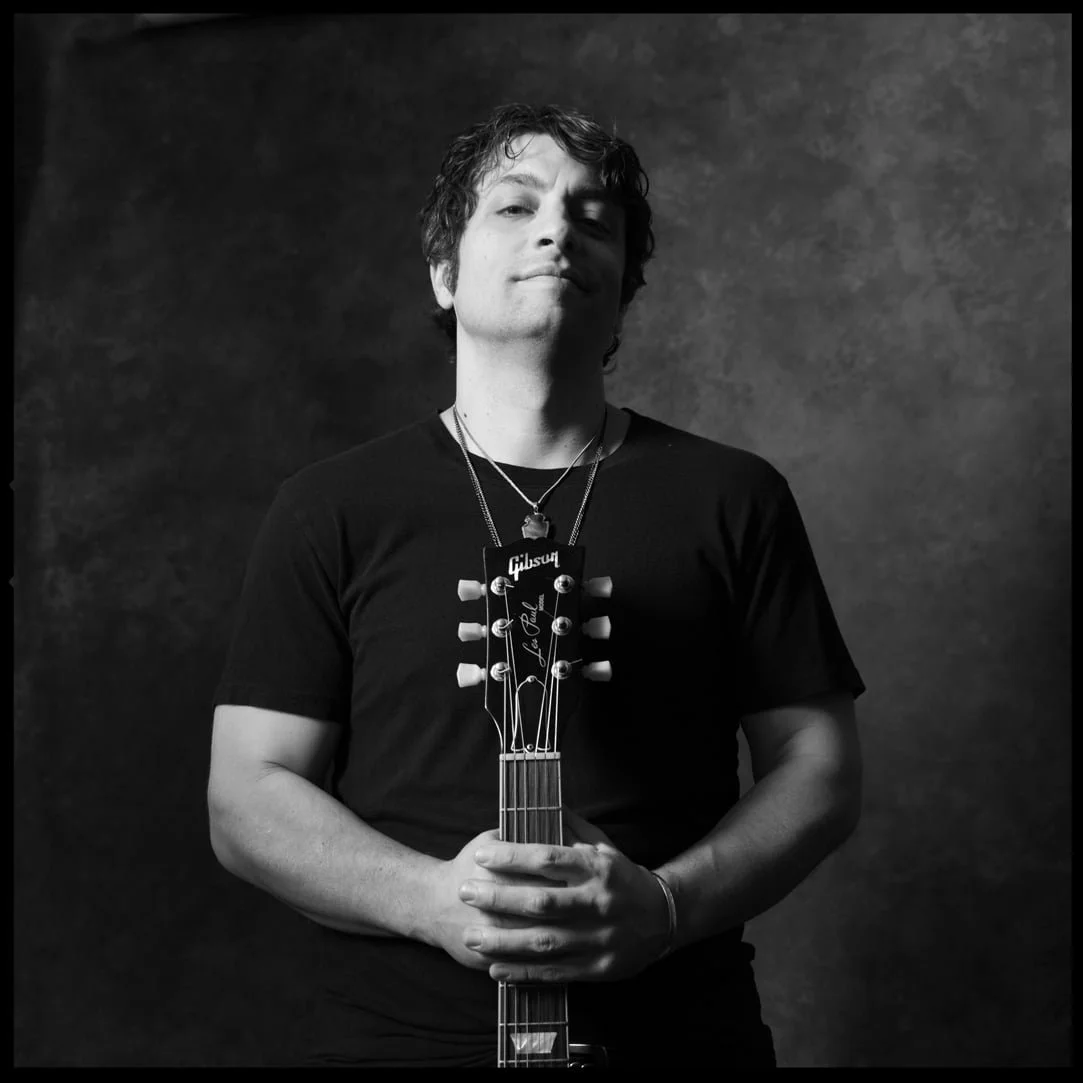PETE MURANO, A man with curly hair wearing a black t-shirt holding an electric guitar, standing against a textured dark background, with a confident expression.