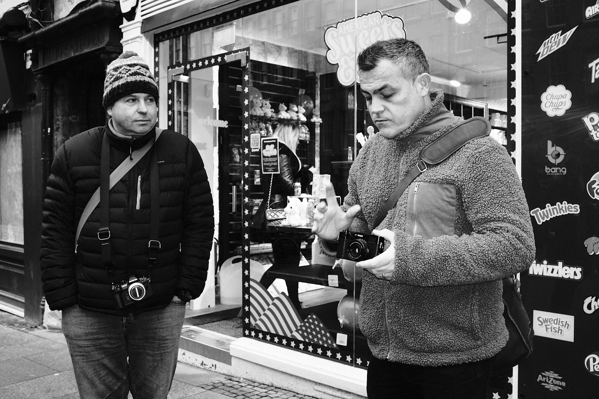 Two men stand outside a store window. One man in a beanie and puffy jacket looks at the other, who is looking at his phone and holding a camera. The store window displays plush toys and signs for candies and snacks.