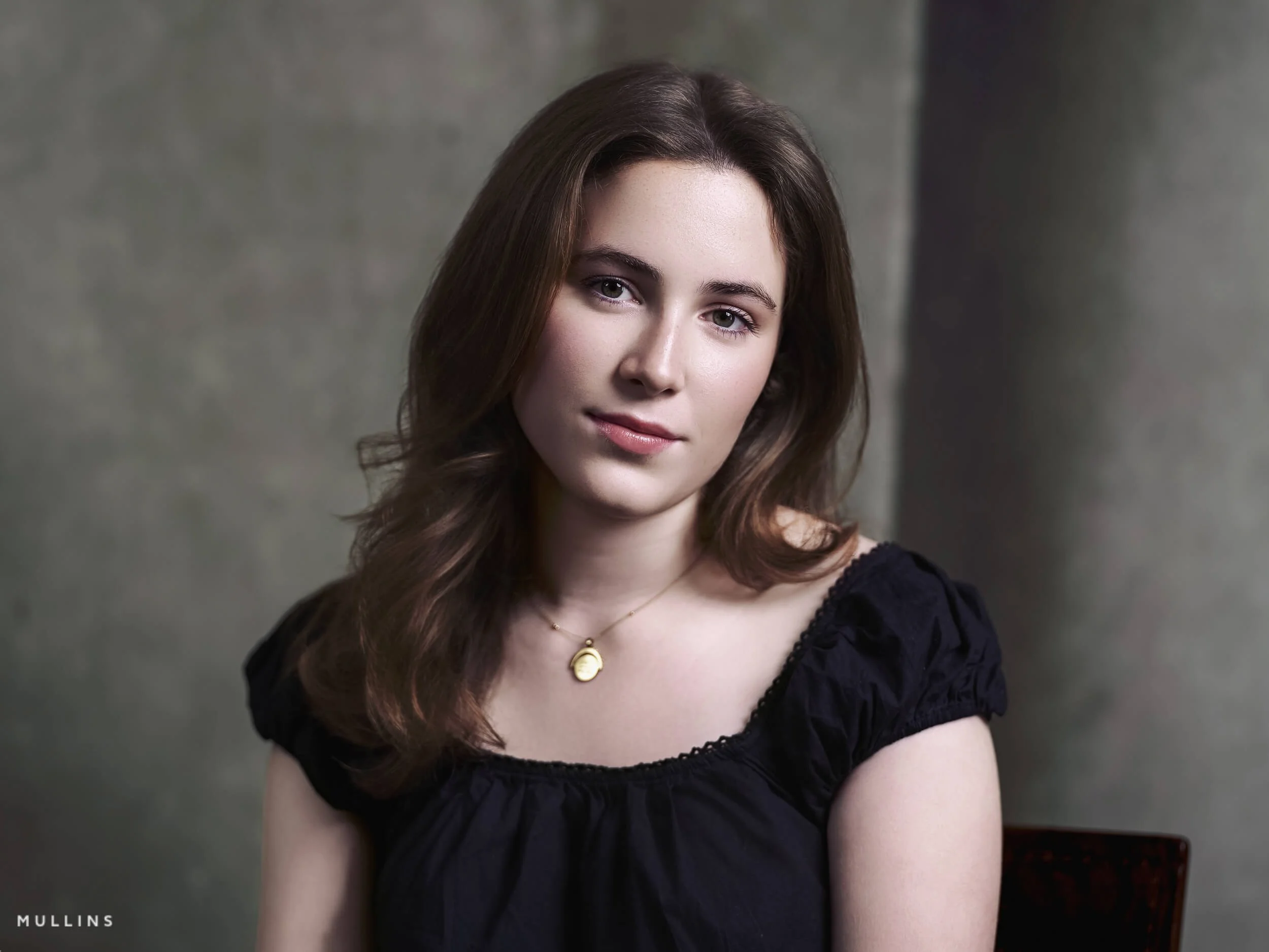Actor portfolio photo of a young actress seated on a wooden chair in a white blouse and jeans against a dark textured studio backdrop