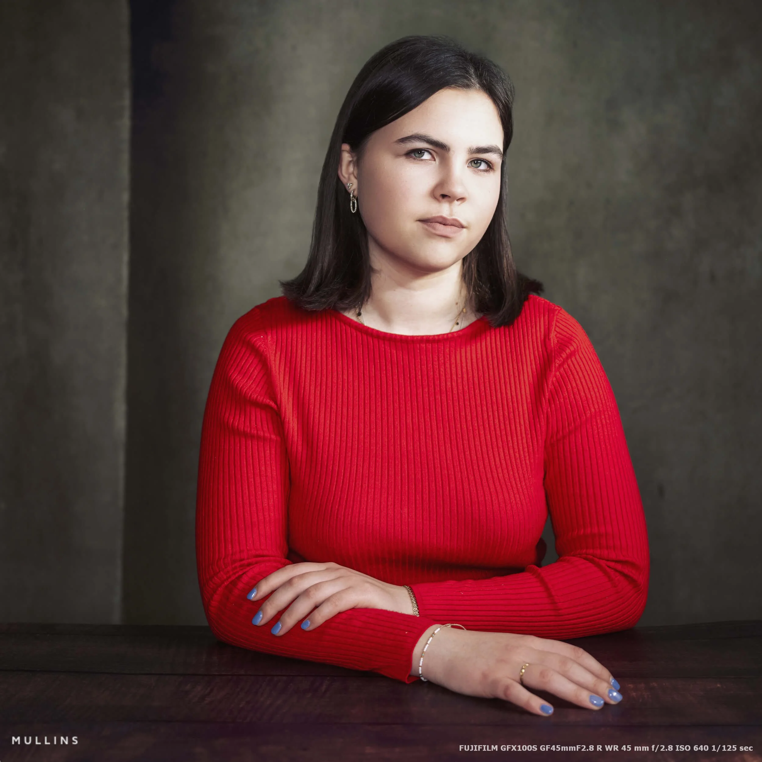 A girl seated at a wooden table in a three-quarter pose, photographed in the studio with soft directional light and a textured backdrop.