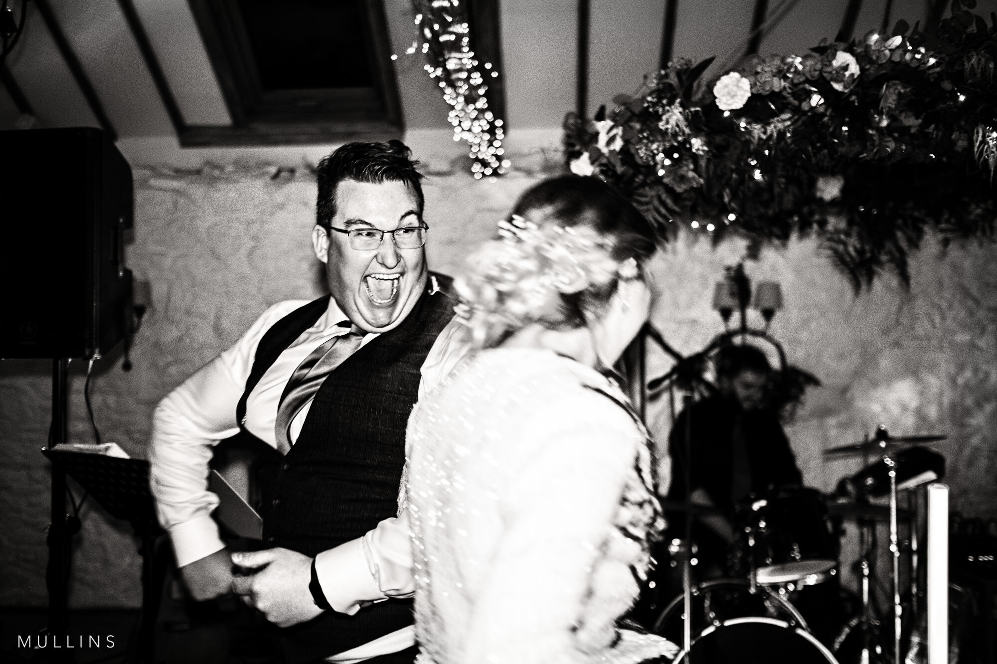 A black and white photo of a joyful man in a tuxedo and glasses dancing at an event with a woman in a sparkly dress and a floral headpiece. The scene includes string lights, floral arrangements, and musical instruments in the background.