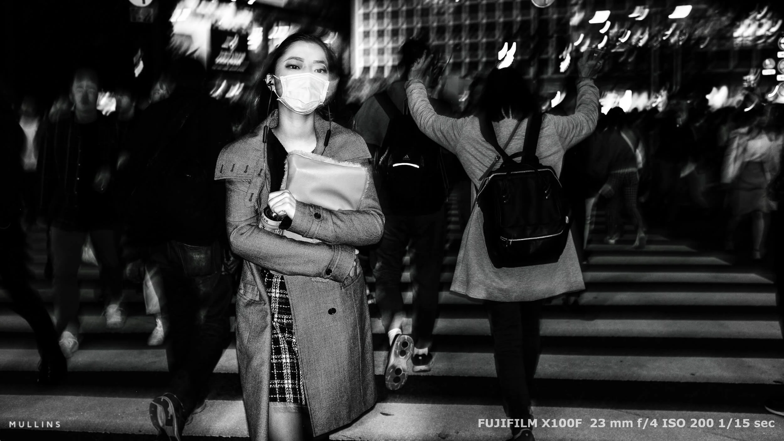 A woman wearing a face mask at a pedestrian crossing, surrounded by blurred figures and streaked background lights.