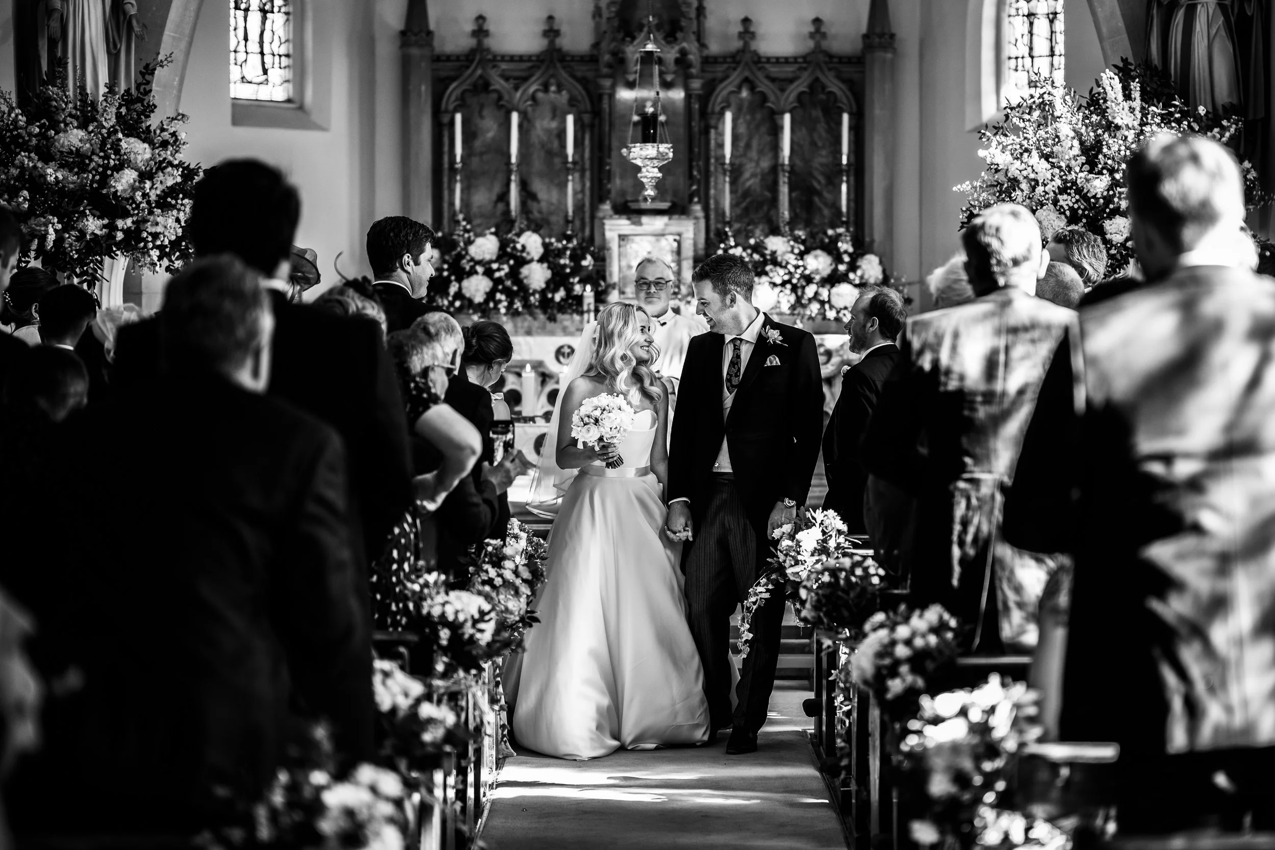 Black and white photo of a wedding ceremony inside a church, with a bride and groom kneeling and holding hands at the altar, surrounded by guests seated along the aisle, decorated with flowers and candles.