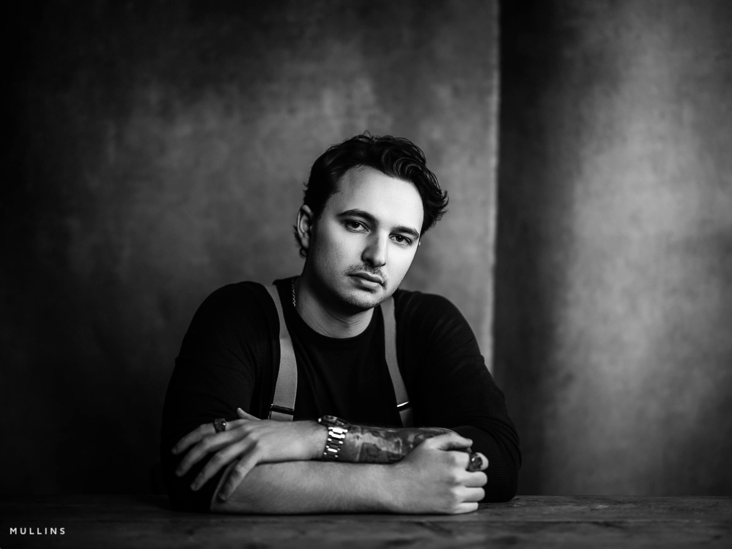 Monochrome business portrait of young entrepreneur leaning on wooden table, wearing dark top and braces in studio.