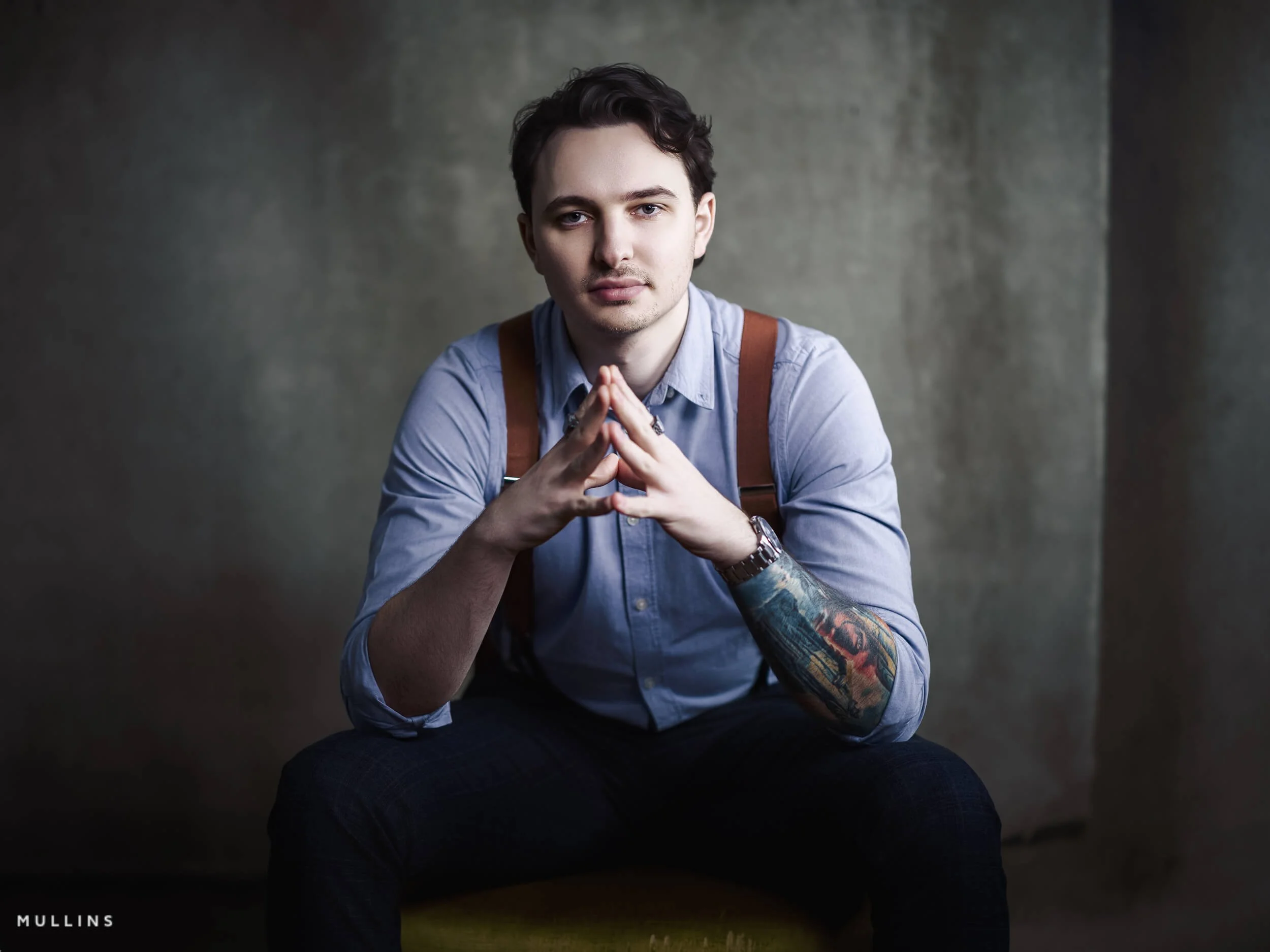 Business branding portrait of young entrepreneur seated forward with hands together, wearing blue shirt and braces against a textured studio backdrop.