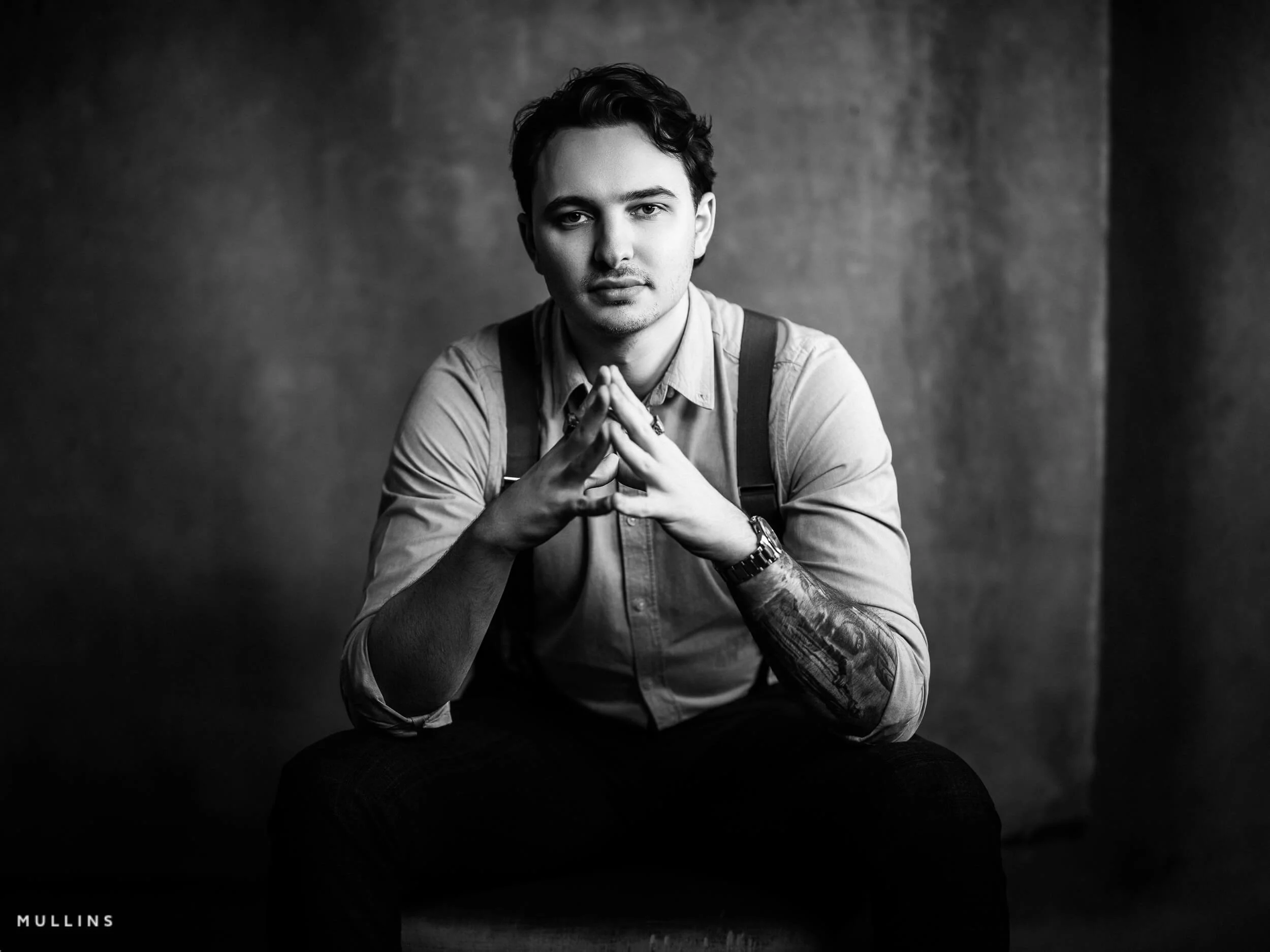 Monochrome branding portrait of young entrepreneur seated forward with hands together, studio portrait with textured backdrop.