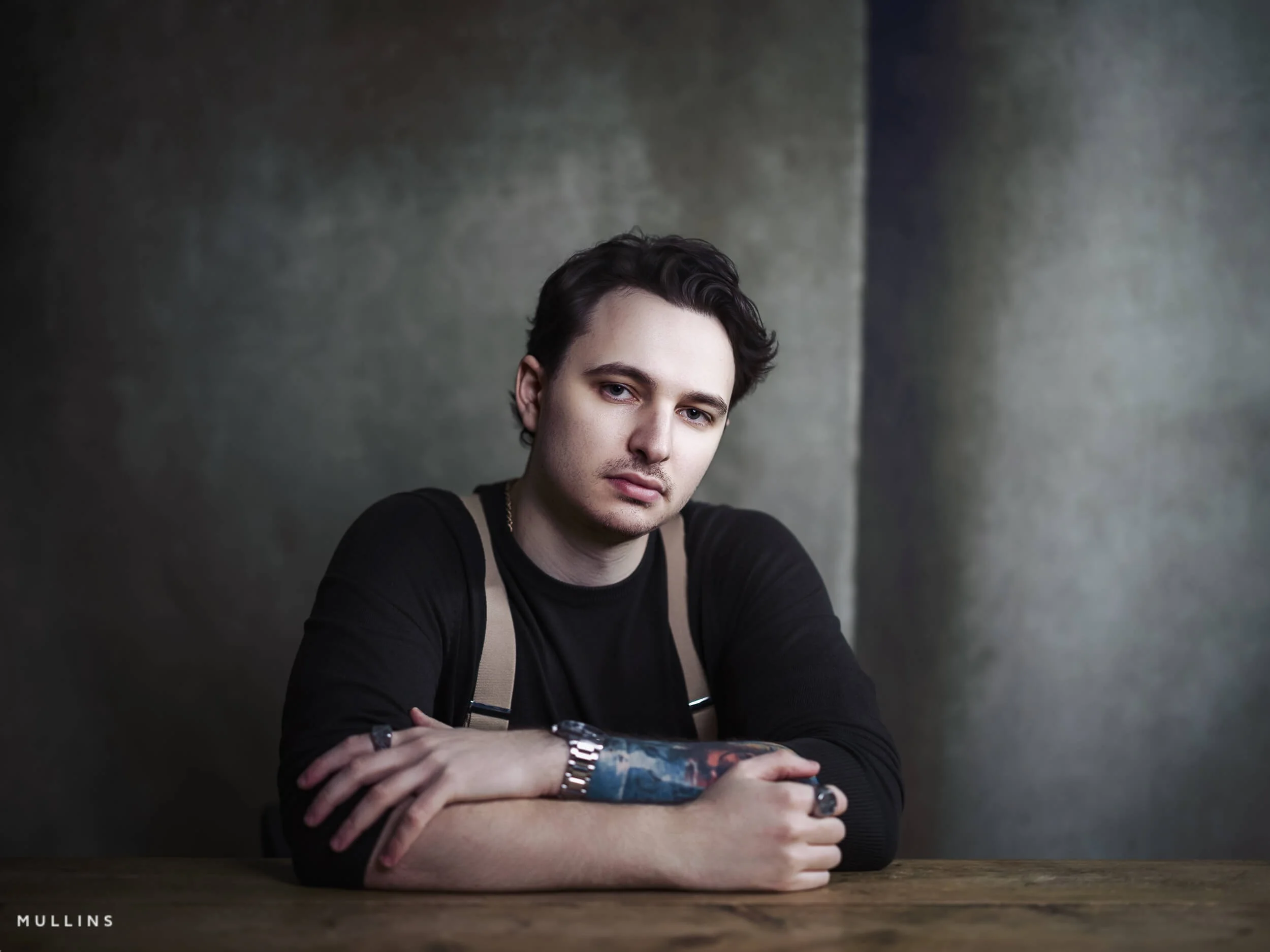 Relaxed business portrait of young entrepreneur leaning on wooden table, wearing dark top and braces, photographed in studio.
