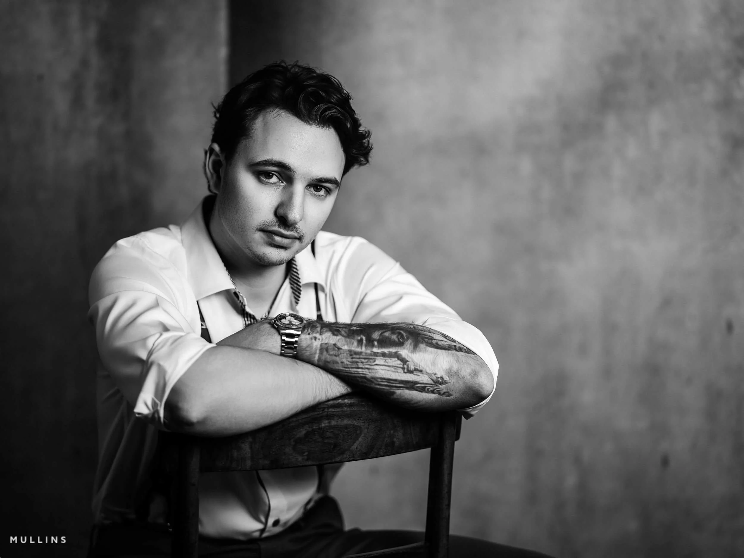 Monochrome studio portrait of young entrepreneur leaning over wooden chair, wearing white shirt and braces.