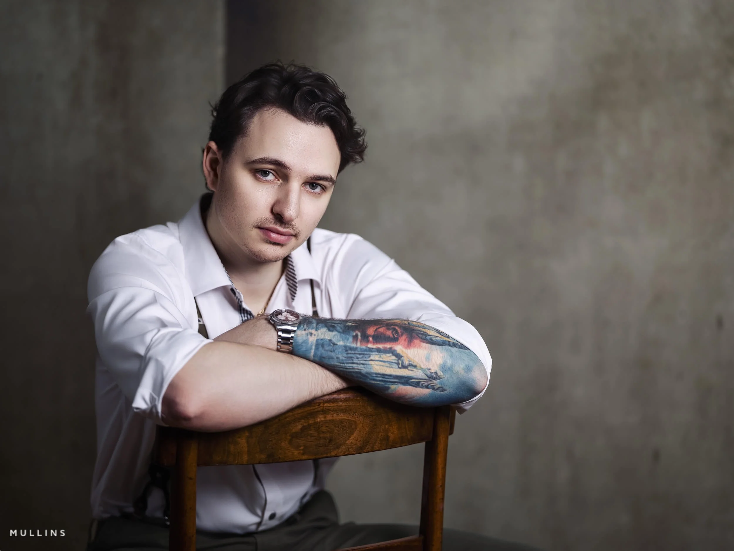 Studio branding portrait of young entrepreneur leaning over wooden chair, wearing white shirt and braces, showing tattooed forearm.