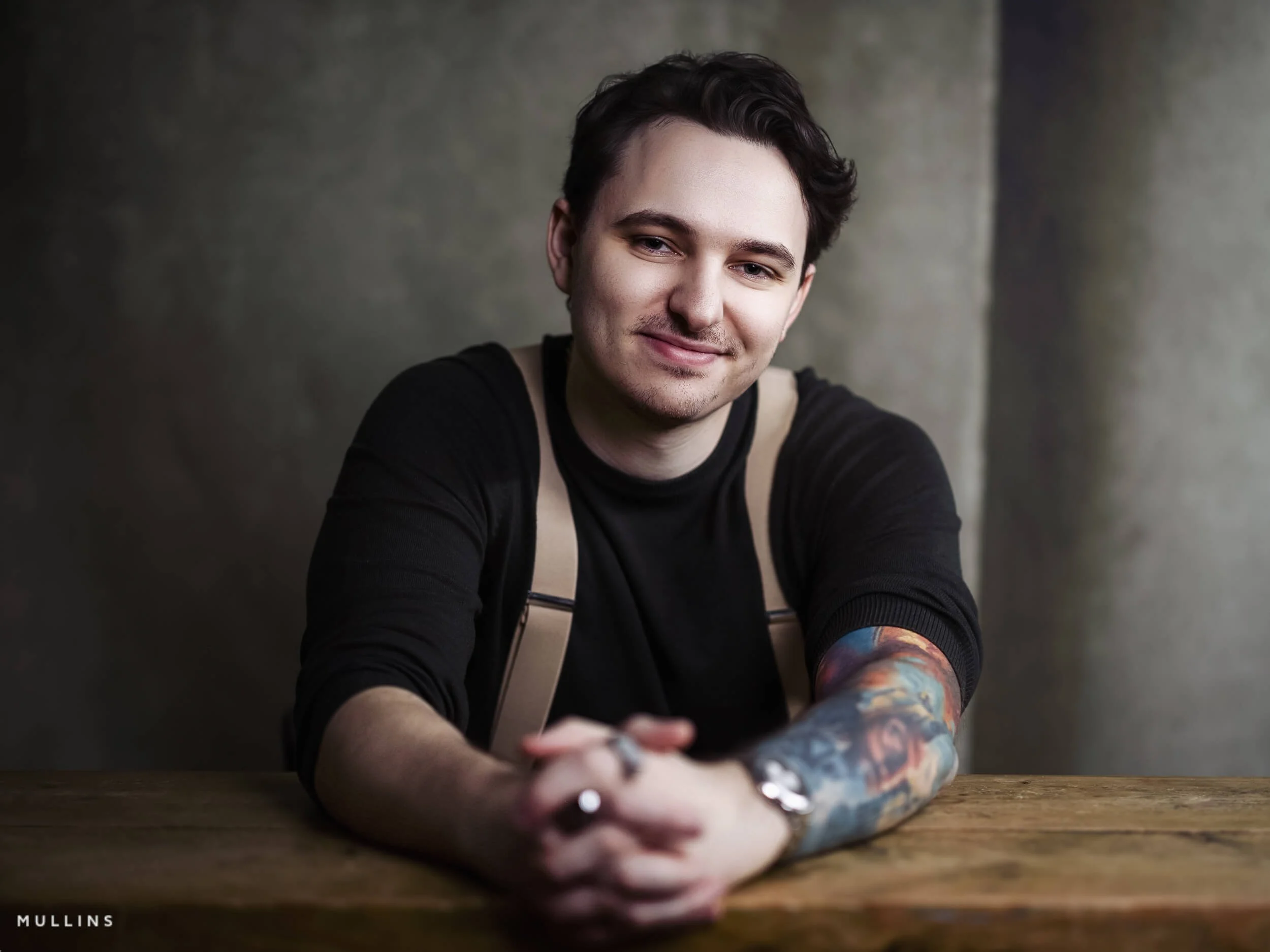 Smiling business branding portrait of young entrepreneur leaning forward on wooden table, wearing dark top and braces.