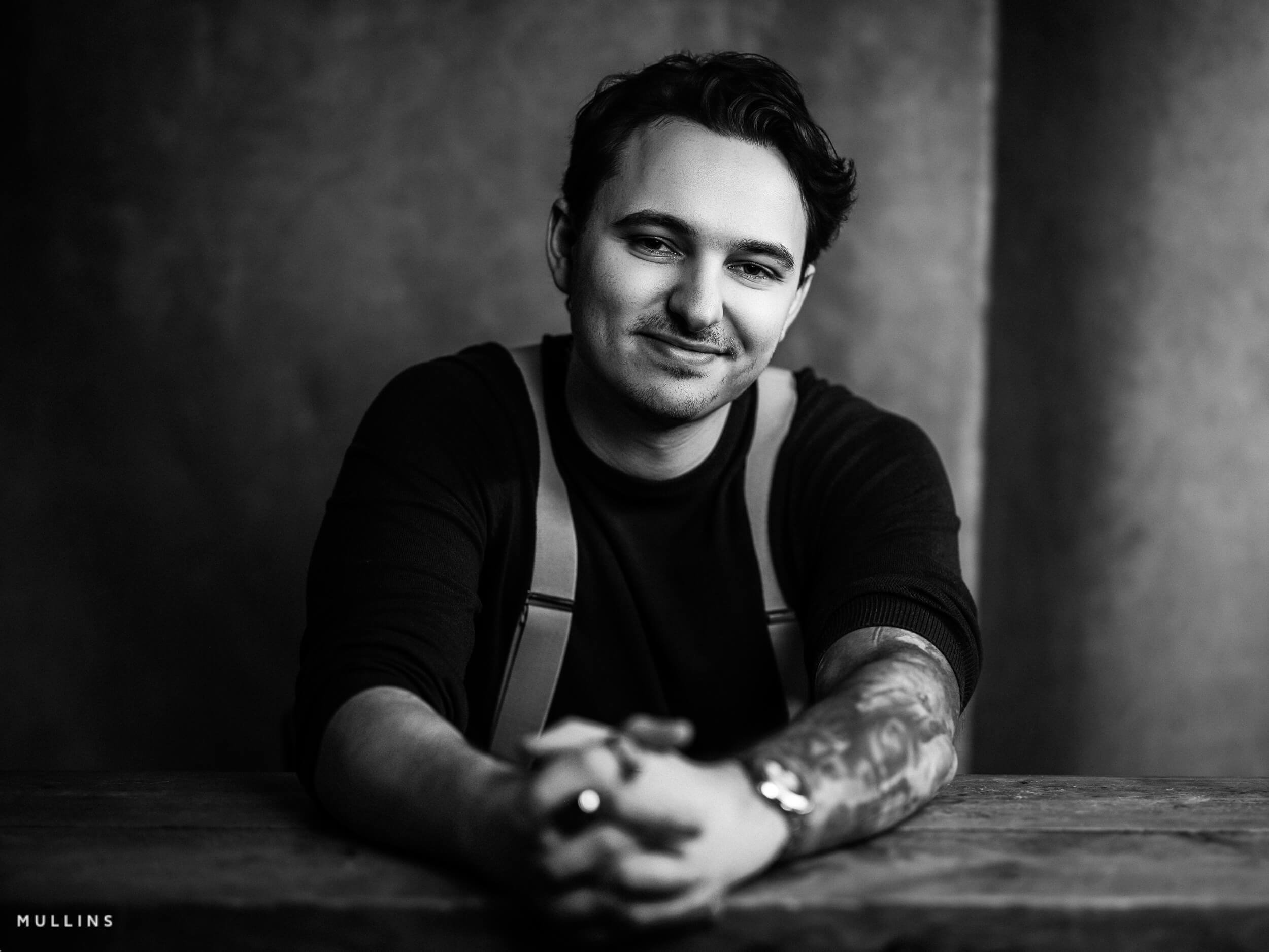Smiling business monochrome branding portrait of young entrepreneur leaning forward on wooden table, wearing dark top and braces.