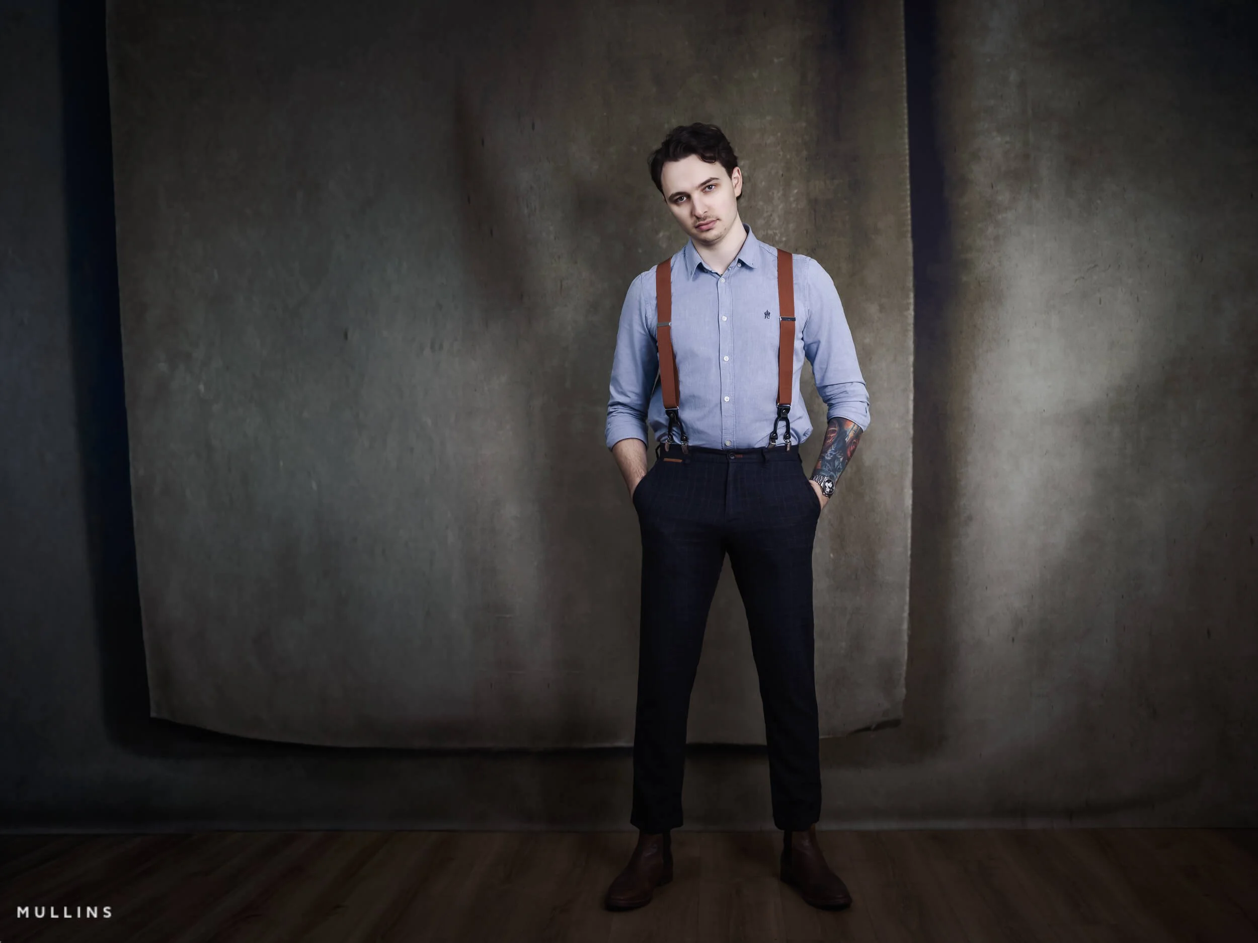 Full-length business branding portrait of young entrepreneur standing in studio wearing blue shirt, braces and tailored trousers.