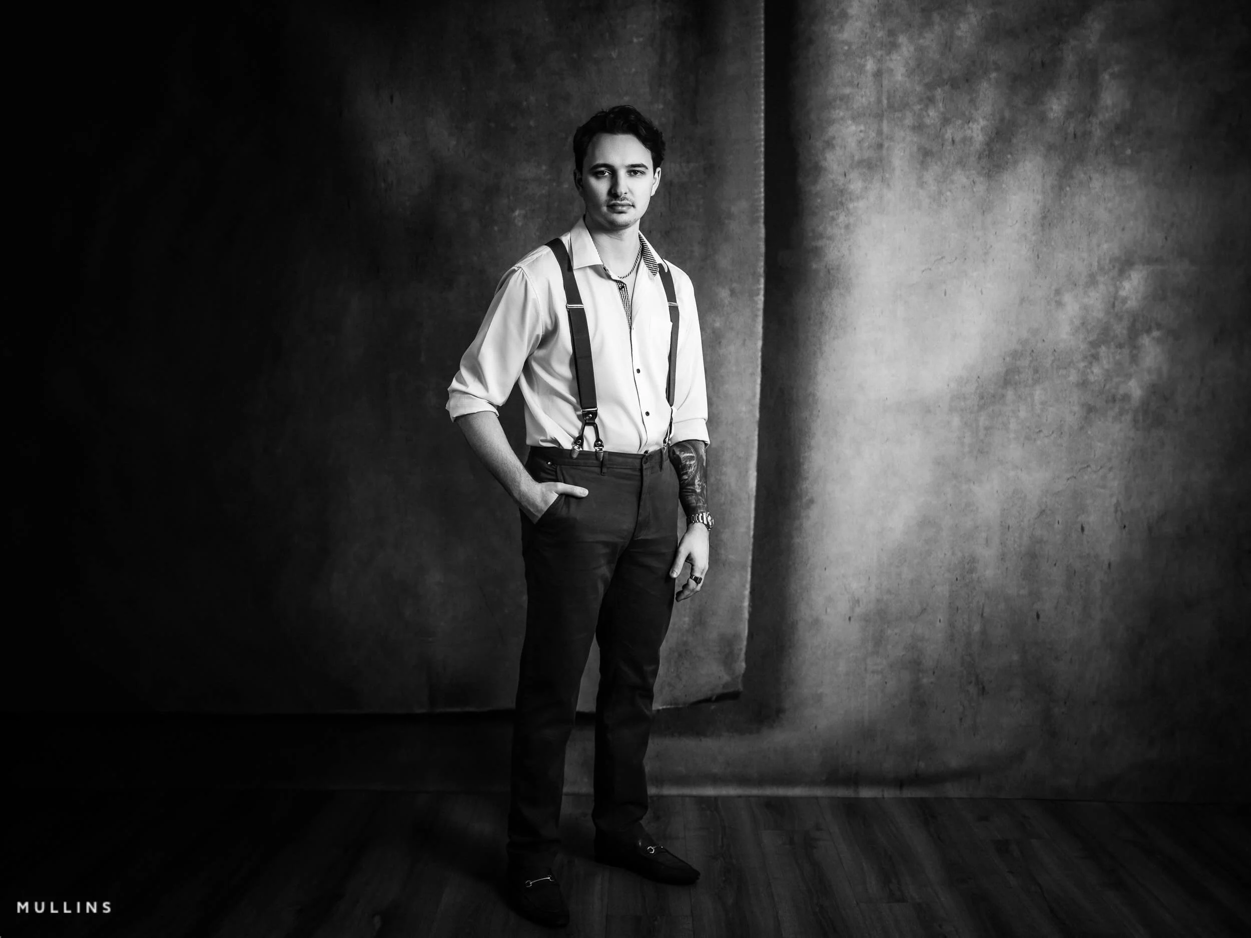 Young businessman standing full length in studio against a textured backdrop, wearing rolled-sleeve shirt and braces, monochrome portrait.