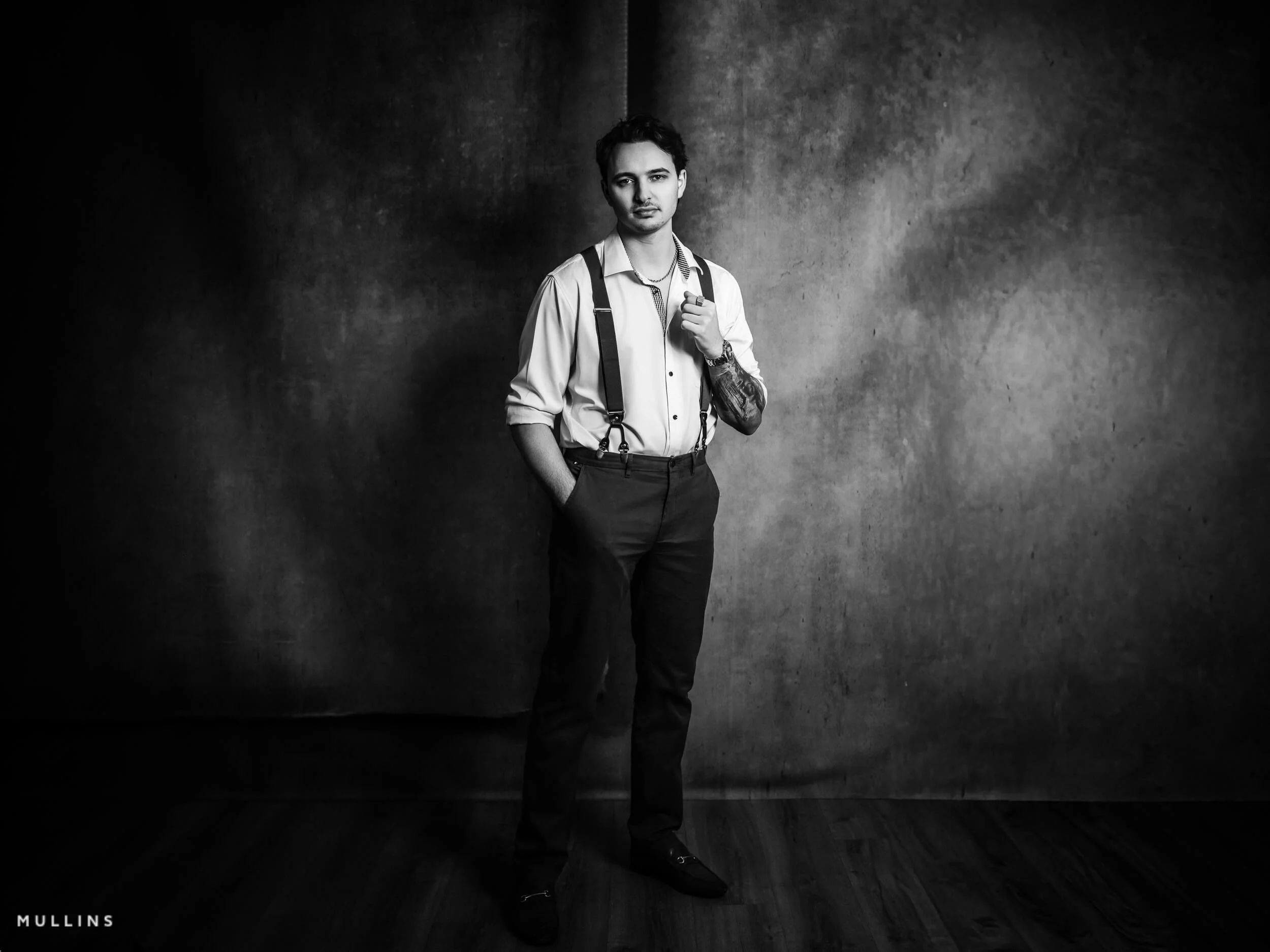 Young entrepreneur standing in studio in white shirt and braces against textured backdrop, business branding portrait.