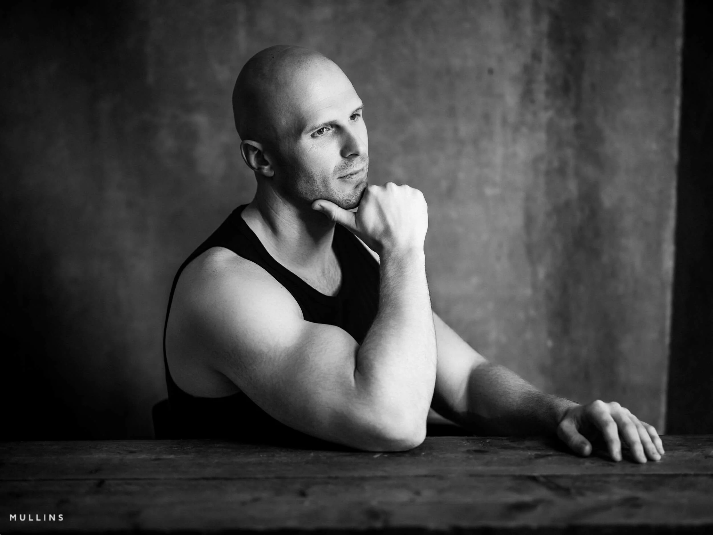 Monochrome side-profile actor portrait of Ian McIntyre seated at a wooden table wearing a black vest.