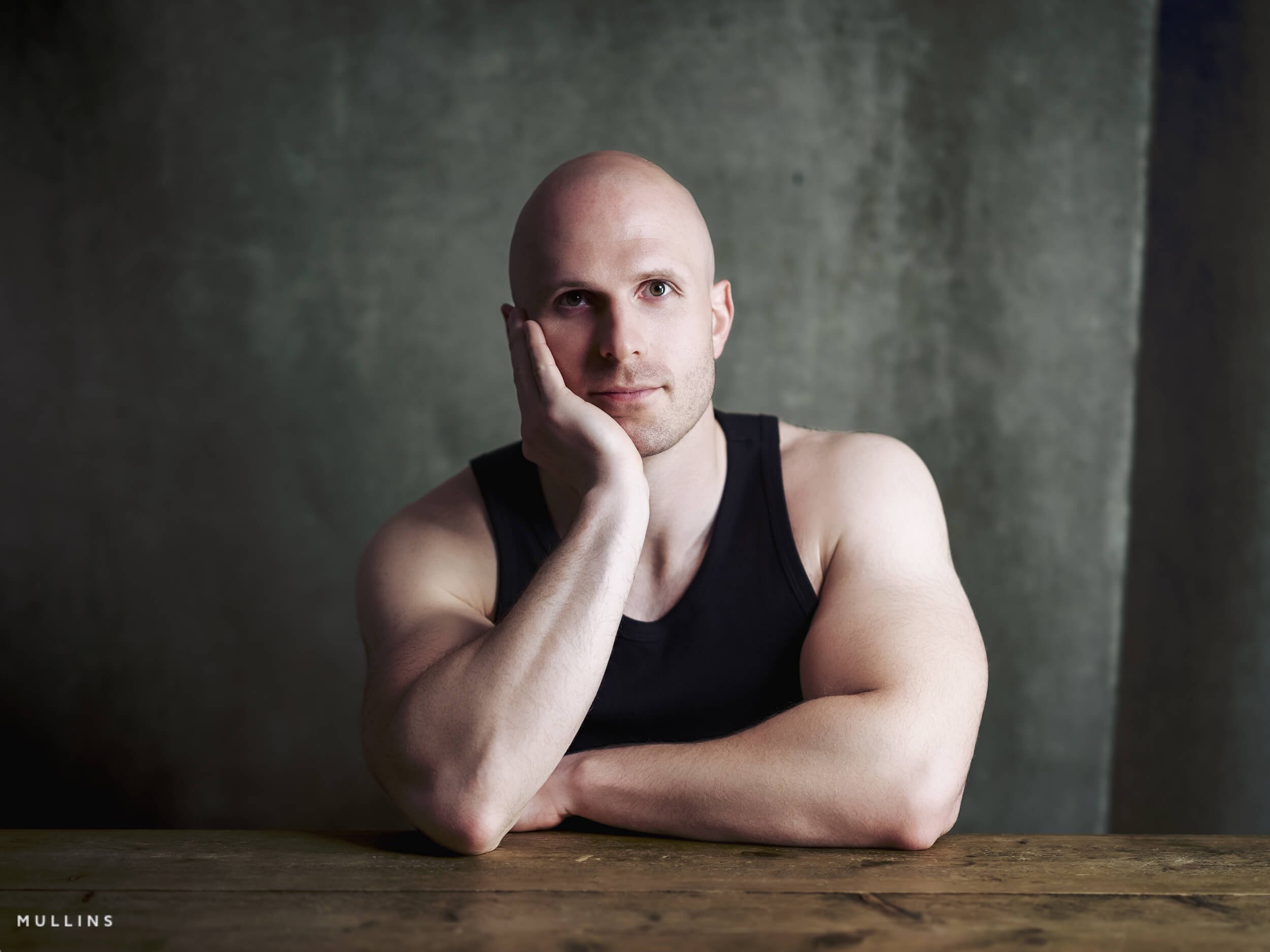Actor portrait of Ian McIntyre seated at a wooden table wearing a black vest, looking directly towards camera.