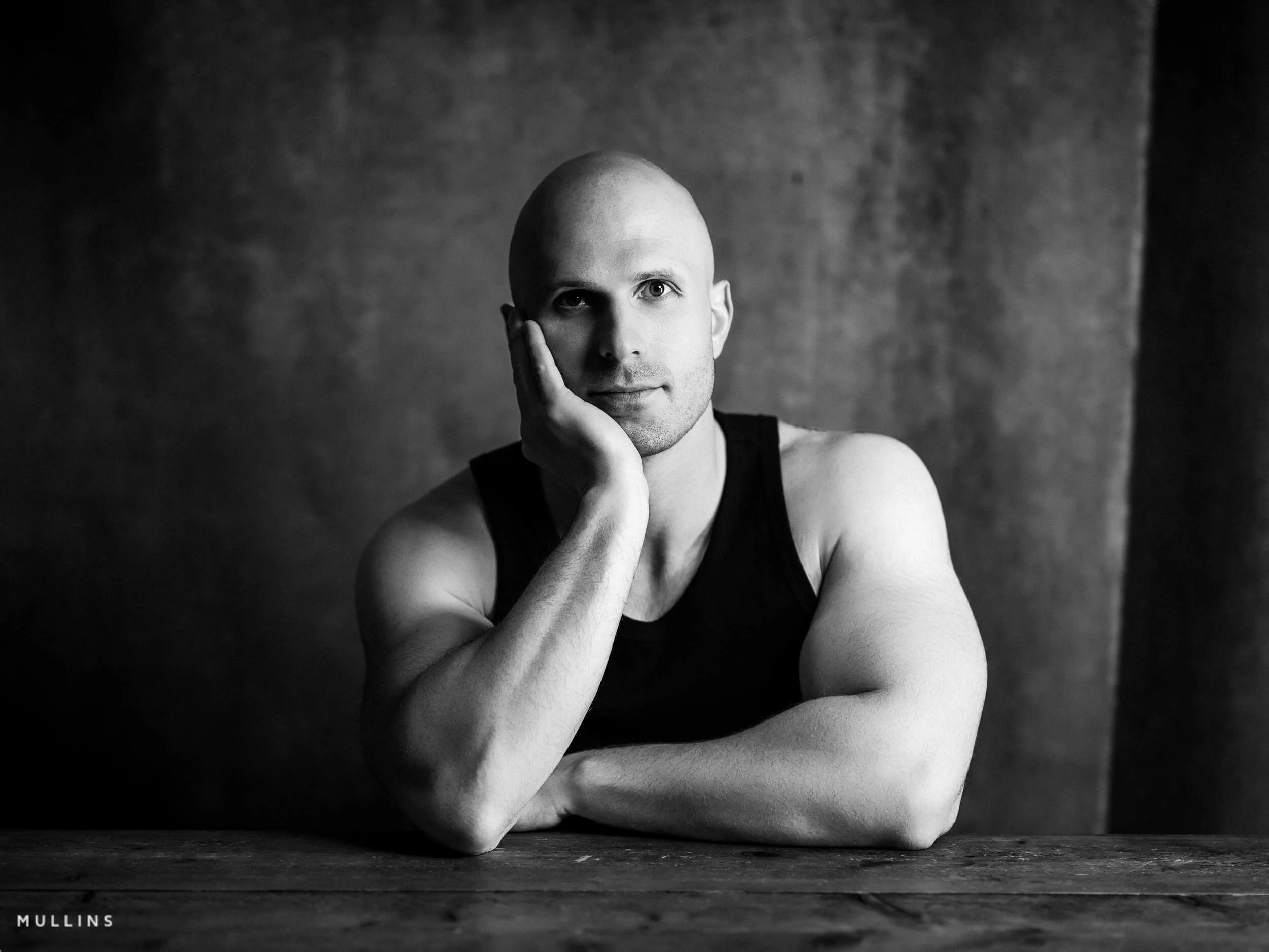 Monochrome actor portrait of Ian McIntyre seated at a wooden table wearing a black vest.