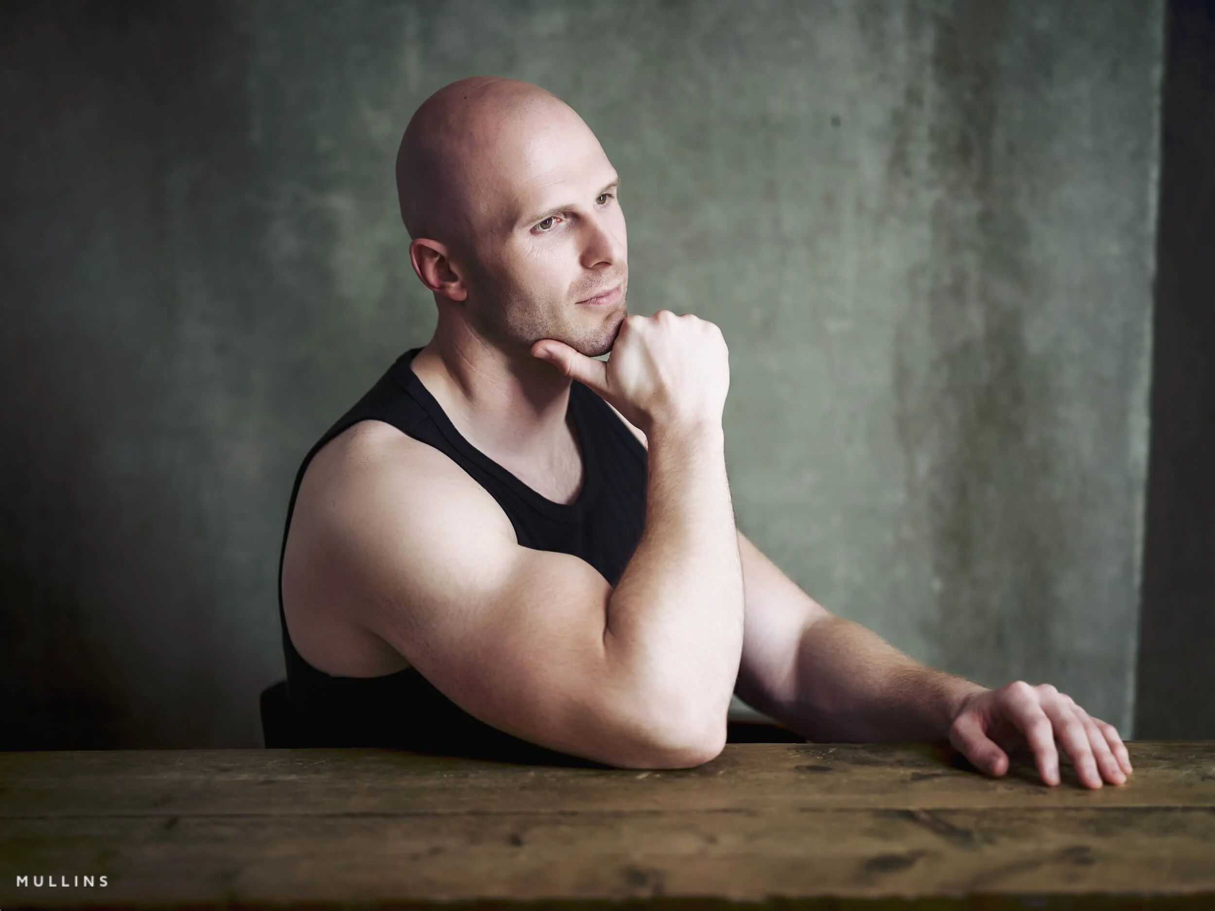 Side-profile actor portrait of Ian McIntyre seated at a wooden table with hand at chin, wearing a black vest.