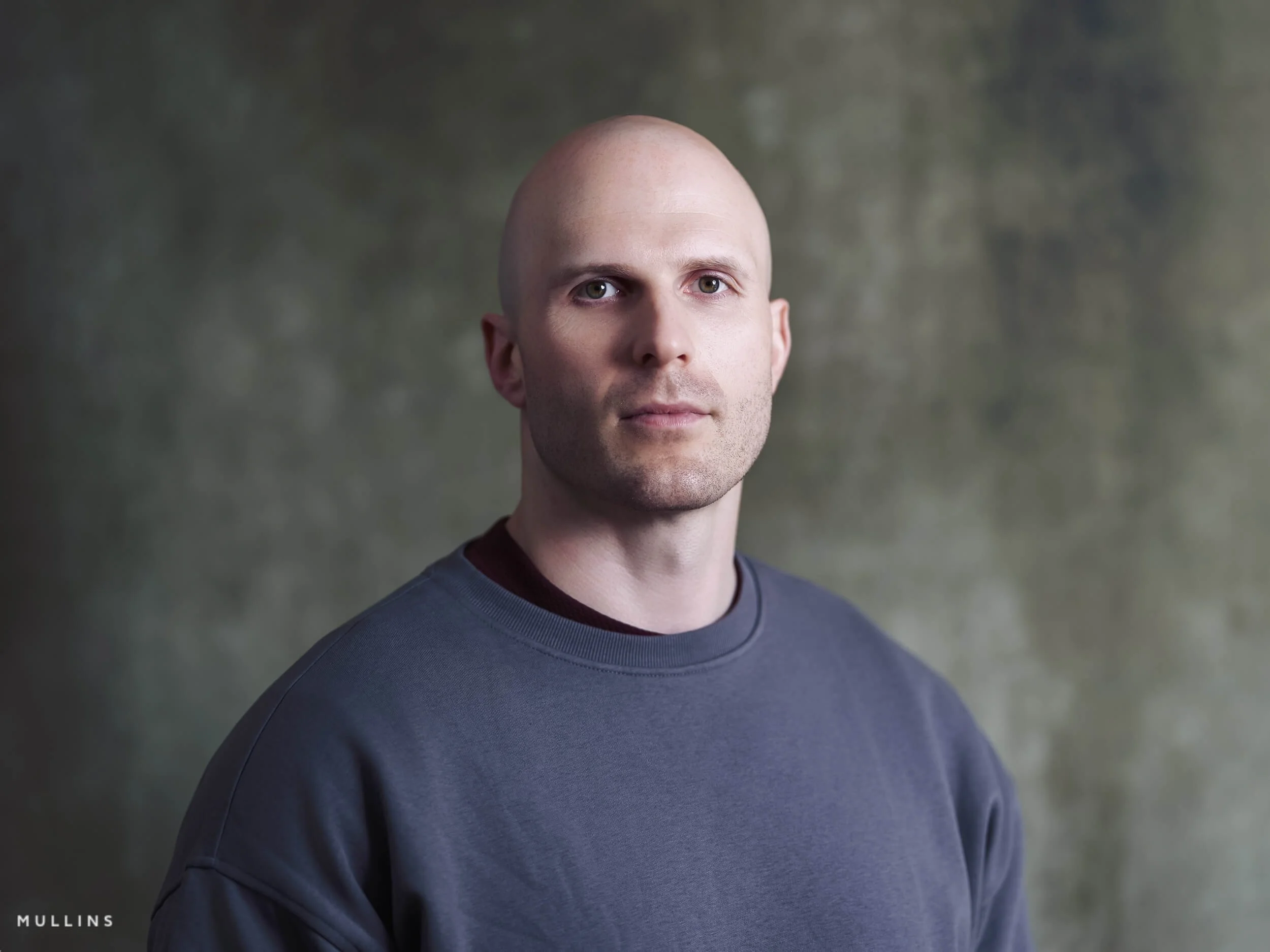 Close-up actor headshot of Ian McIntyre in studio wearing a grey sweatshirt against a soft textured background.