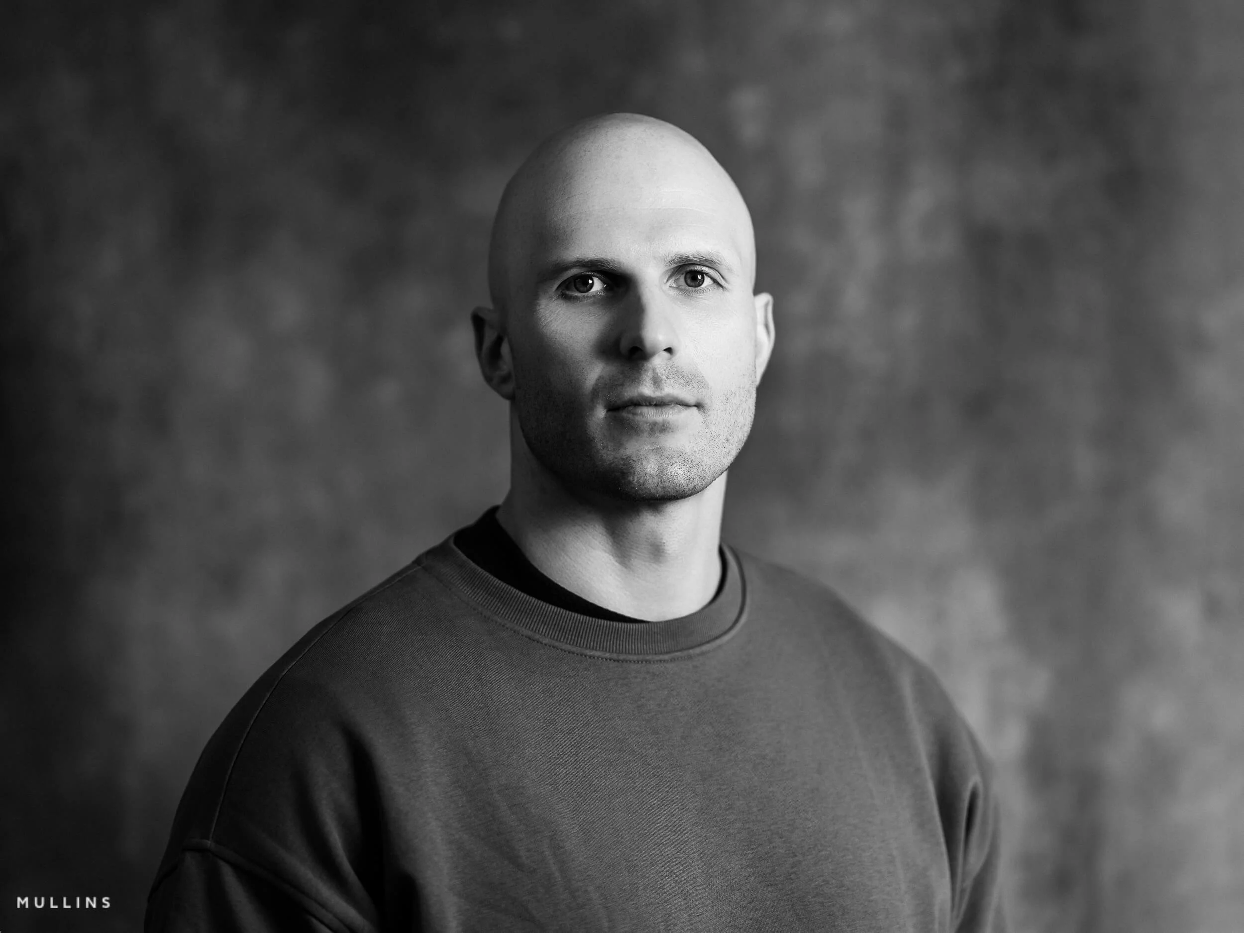 Monochrome actor headshot of Ian McIntyre wearing a grey sweatshirt in studio against a textured backdrop.