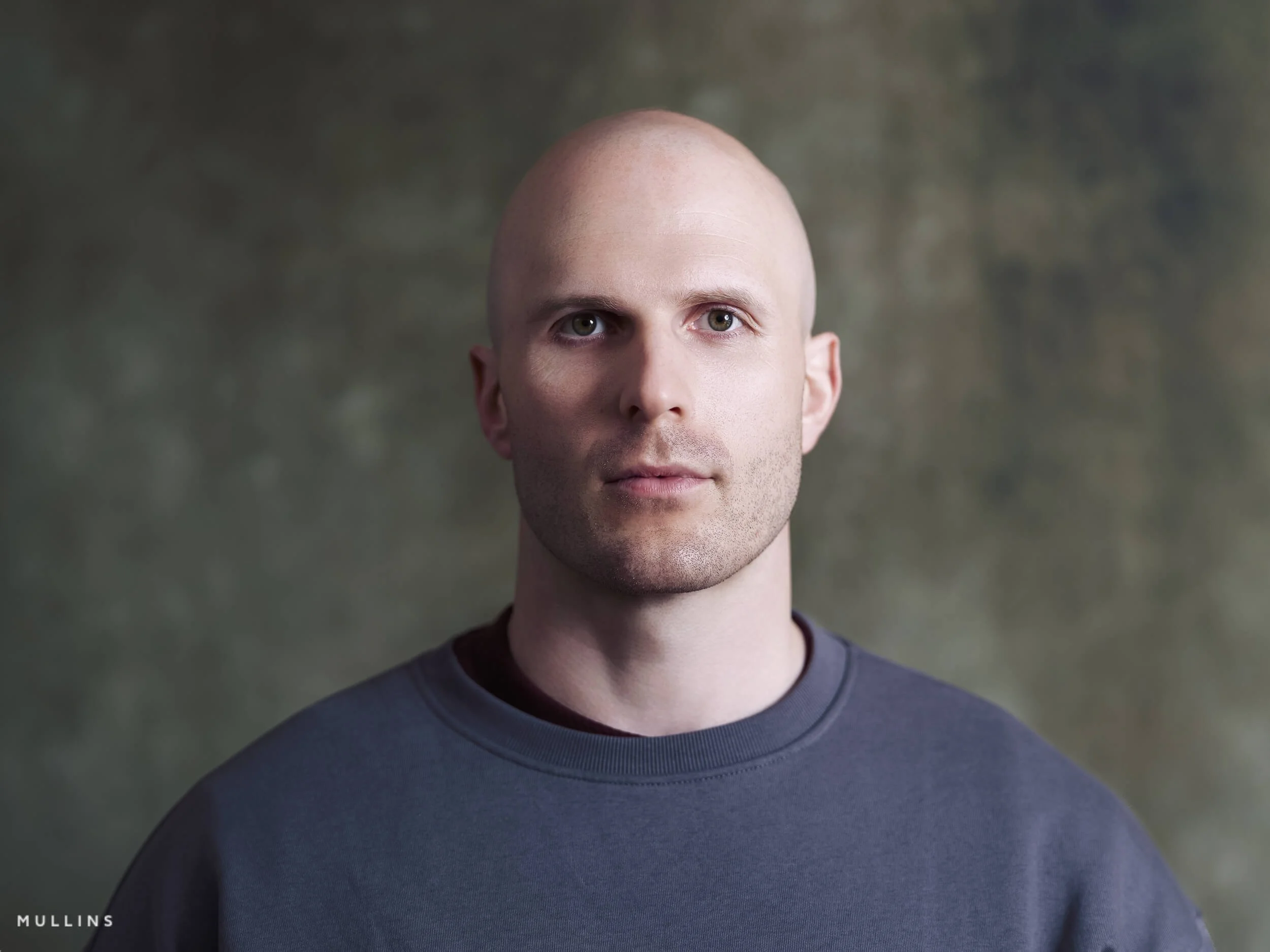Front-facing actor headshot of Ian McIntyre in studio wearing a grey sweatshirt against a soft textured background.