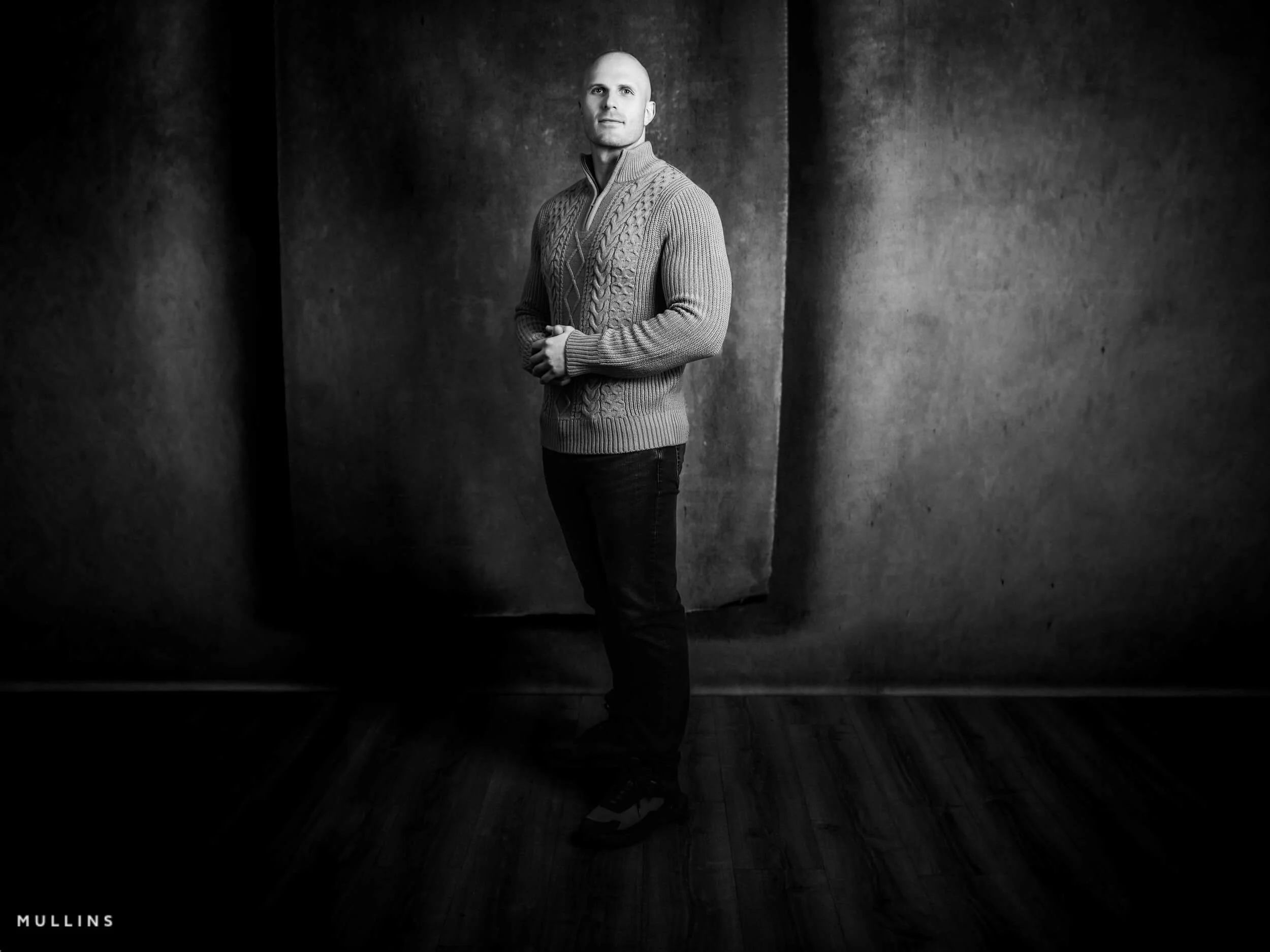 Full-length monochrome portrait of actor Ian McIntyre standing in studio wearing a zip-neck knit jumper against a textured backdrop.