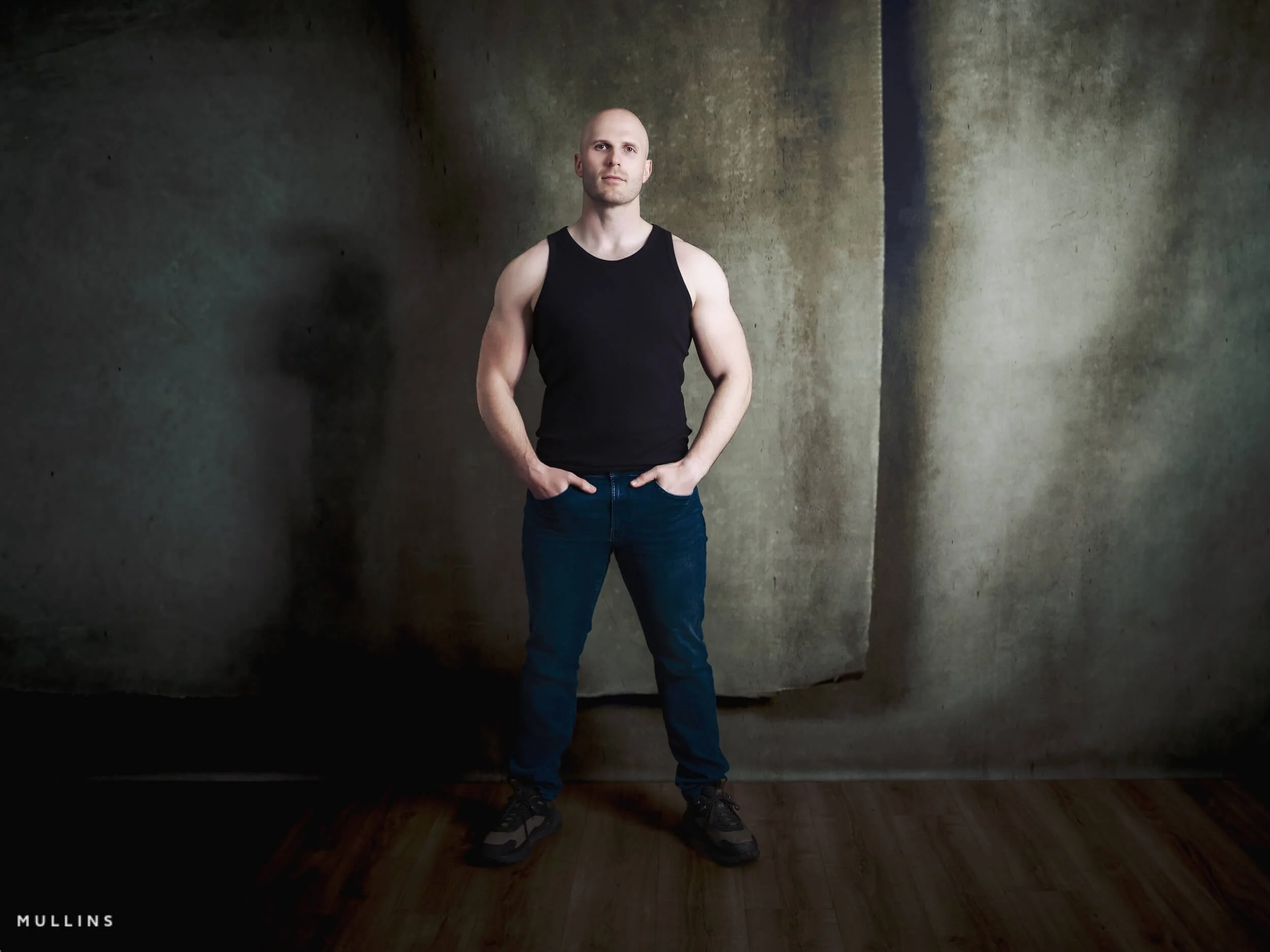 Full-length actor portrait of Ian McIntyre standing in studio wearing a black vest and jeans against a textured backdrop.