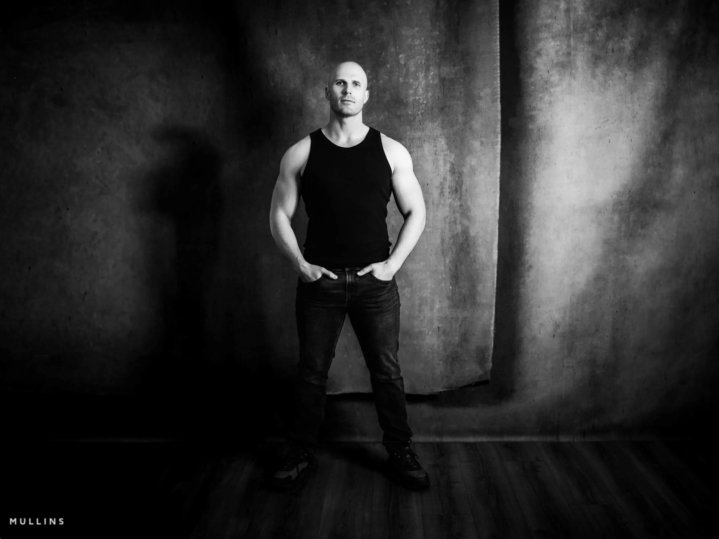 Full-length monochrome actor portrait of Ian McIntyre standing in studio wearing a black vest and jeans.