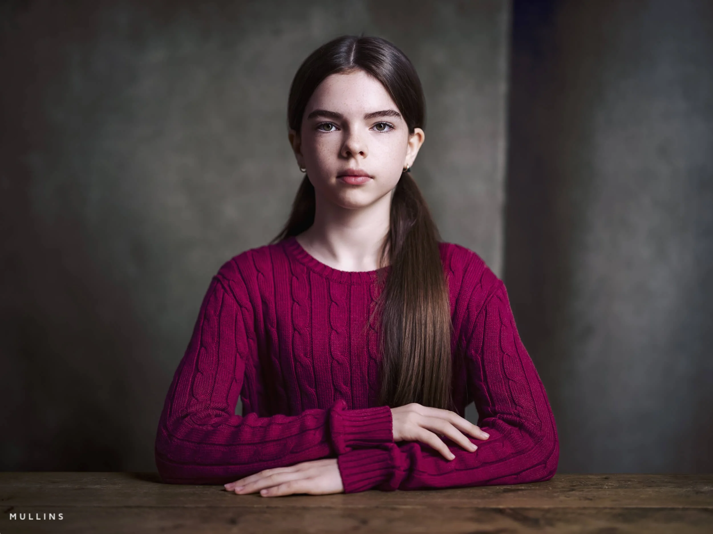 ortrait of a young female actor seated at a wooden table with a calm expression, wearing a bright jumper.
