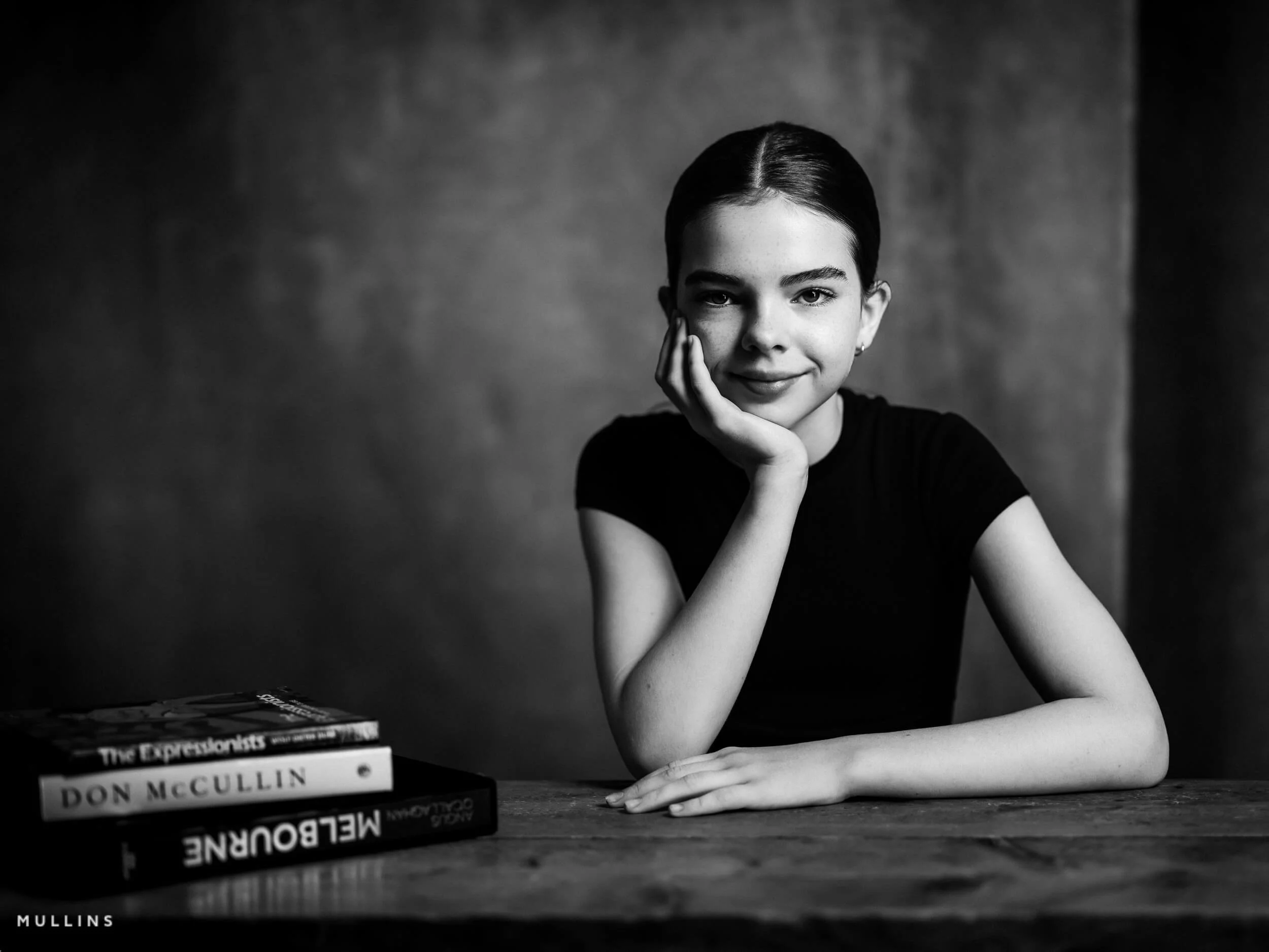 Black and white portrait of a smiling young female actor seated at a wooden table with books beside her.
