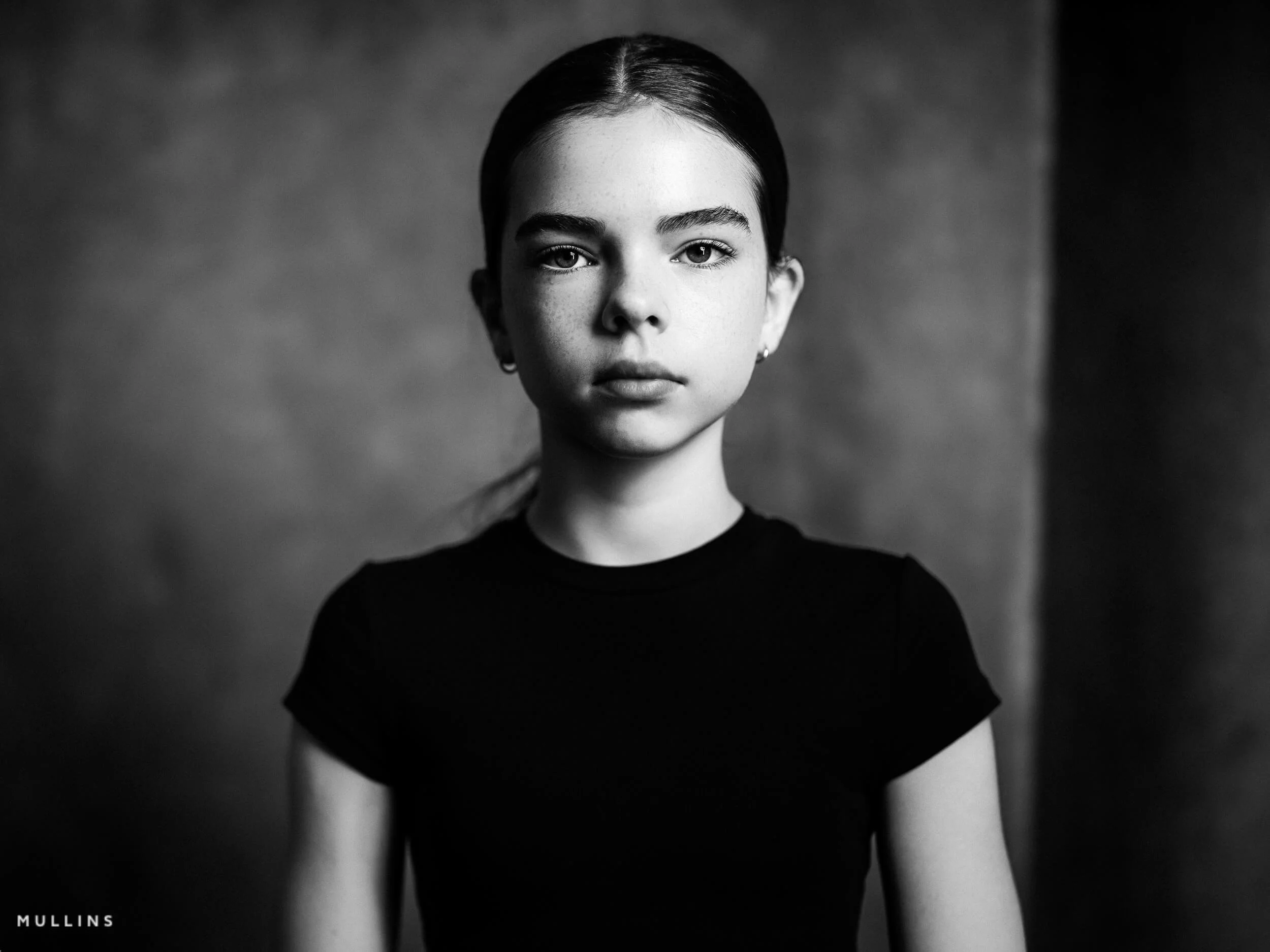 Black and white close portrait of a young female actor wearing a black top against a textured studio backdrop.